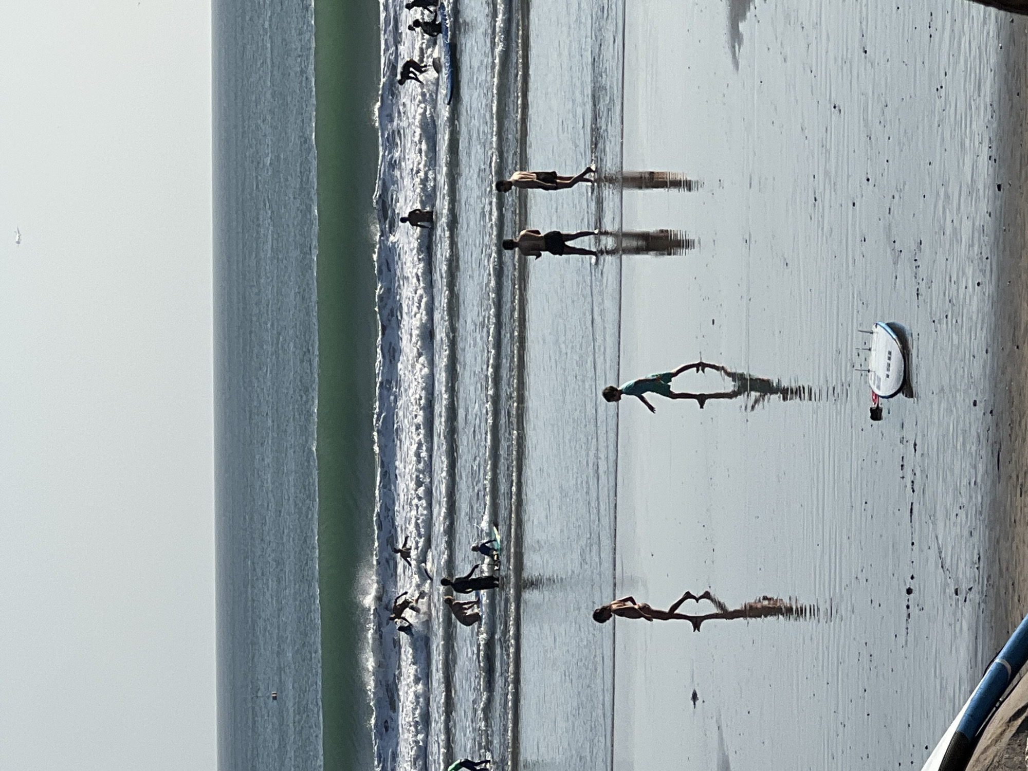 The image depicts a crowded beach in Kuta, Indonesia, with many individuals engaged in various activities in and around the ocean. The scene is characterized by a large, green wave breaking in the middle ground, with numerous surfers attempting to ride it. Several people are in the water, some on surfboards, while others are swimming or wading. The foreground shows a sandy beach with reflections of the people and the water. Two young children are walking on the wet sand in the foreground, their reflections clearly visible. Further back, a group of men are standing in the shallow water. A small, white boat with blue trim lies on the wet sand in the lower right portion of the image. The sky is overcast, and an airplane is visible in the distance. The overall atmosphere is one of leisure and recreation on a sunny day.