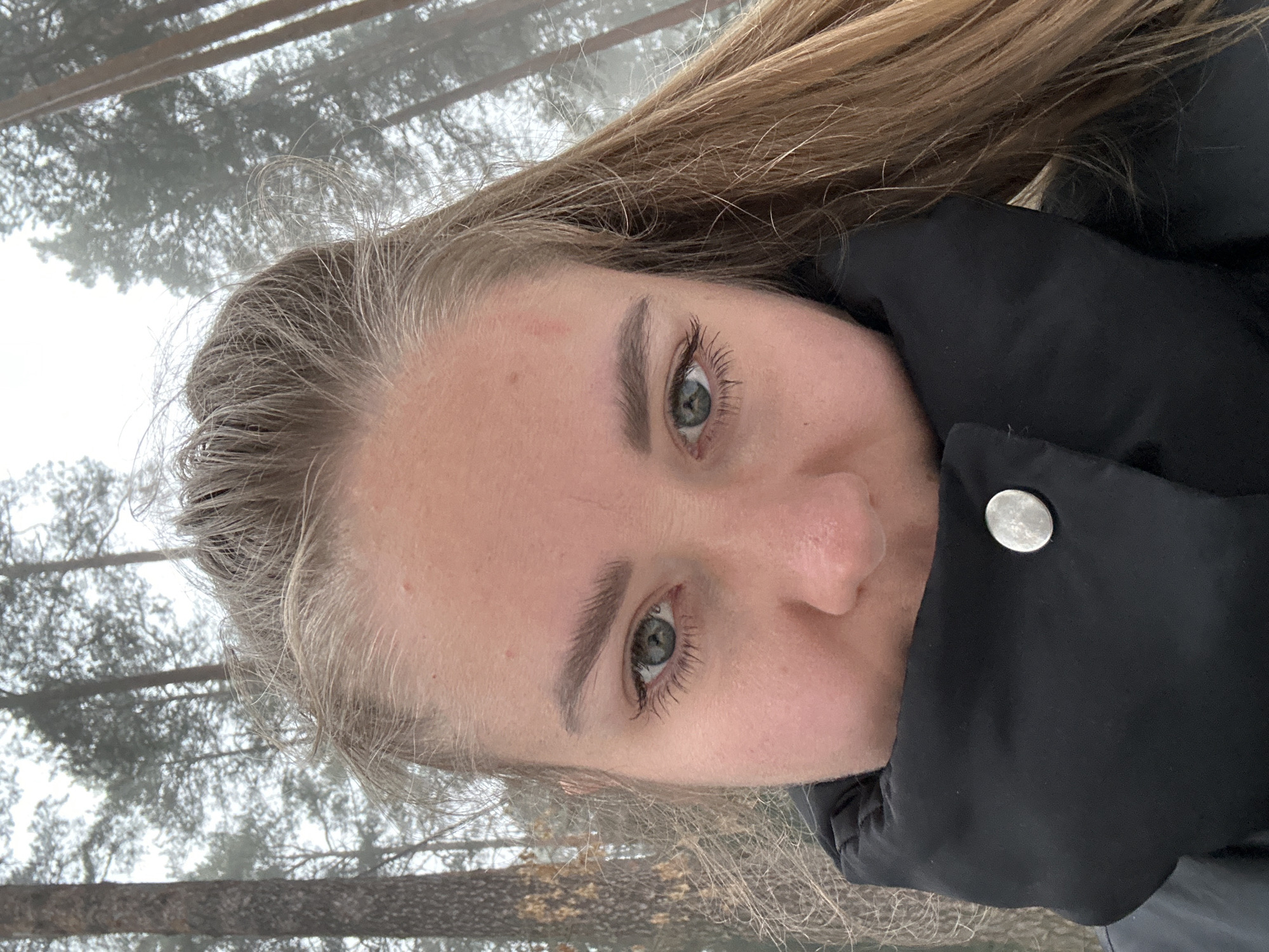 A selfie taken outdoors in a snowy, forested area, likely in a park or wooded area in winter. The individual is a fair-skinned person with blonde hair and green eyes. They are wearing a black puffy jacket with a silver snap button visible. The background includes snow-covered trees and a blurred, overcast sky. It is impossible to confirm the location as Chaikovsky, Russia from the given image.