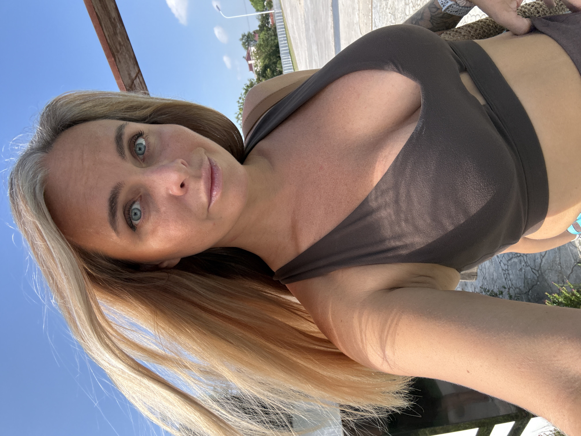A woman with blonde hair and blue eyes is taking a selfie outdoors on a sunny day. She is wearing a brown sports bra. The background shows a paved area, some vegetation, a white fence, a street light, and buildings in the distance. The image appears to be taken in Pudong, China.