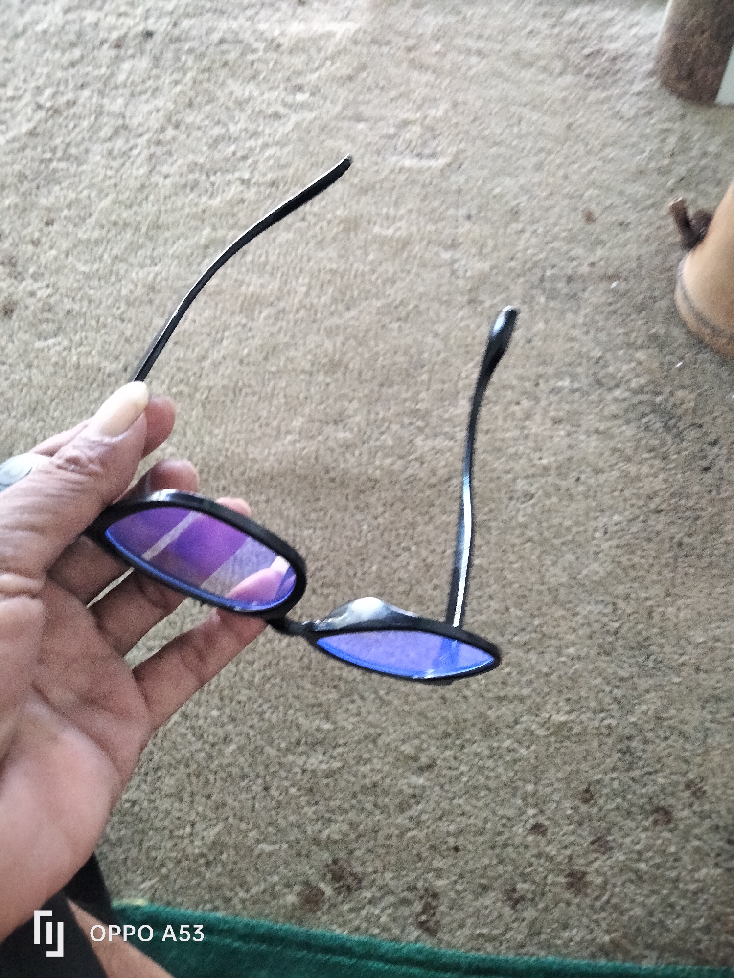 In Badung, Indonesia, a hand holds a pair of black-rimmed sunglasses with purple-tinted lenses. The sunglasses are angled, with one lens facing upwards and the other downwards. The hand is positioned to the left of the frame, with fingers curled around the left temple of the sunglasses. The background consists of a textured, carpet-like surface, possibly a floor or rug, with a muted brown and beige color palette. A dark green fabric is visible at the bottom edge of the image. The lighting suggests an indoor setting, with some reflections visible on the sunglass lenses.