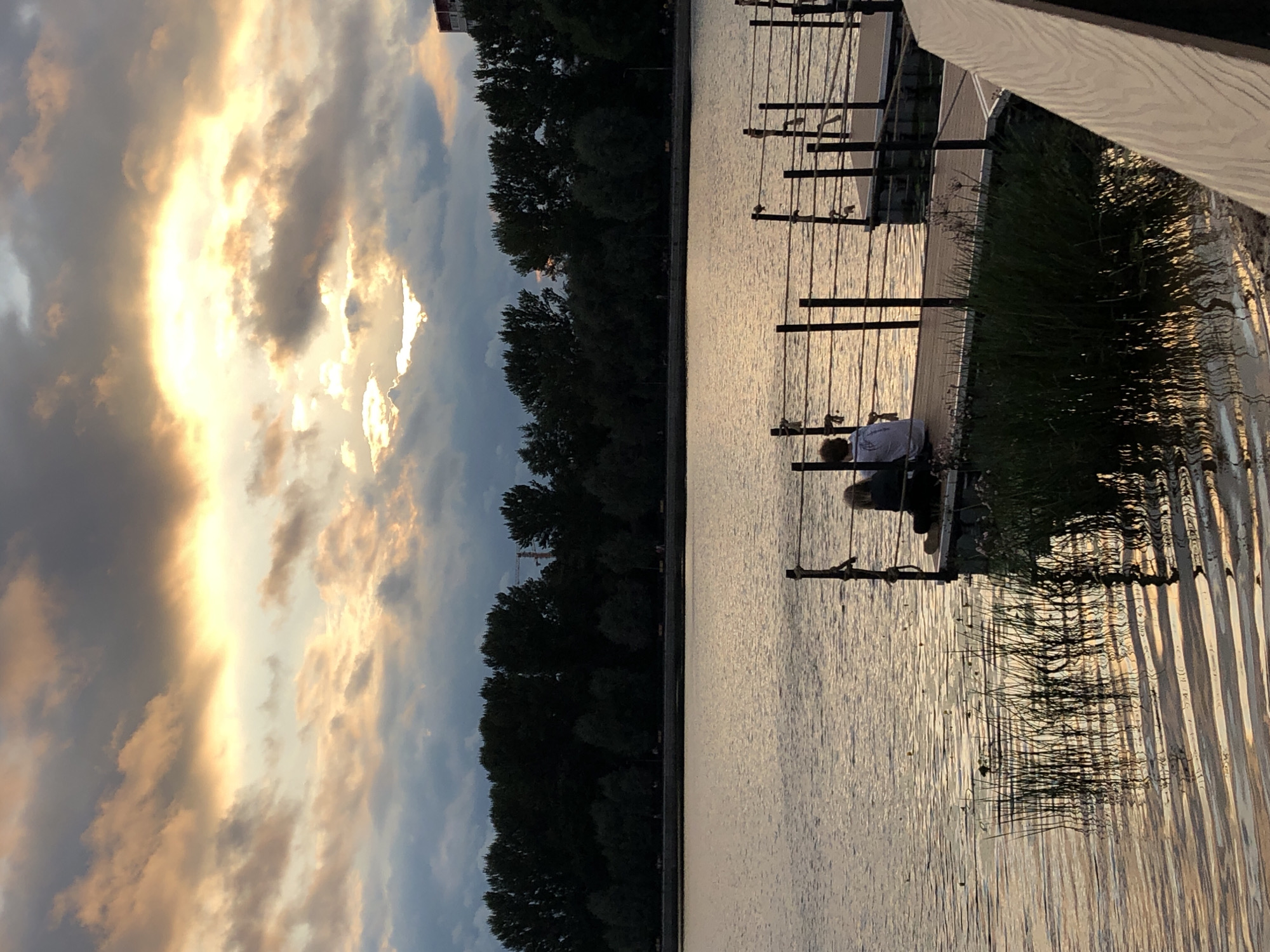 The image shows a sunset over a body of water, likely a lake or river in the city of Khimki, Russia. The sky is filled with dramatic clouds tinged with orange and yellow from the setting sun, with a bright patch of light breaking through. In the foreground, two people are sitting on a wooden dock, facing the water. The dock is surrounded by the calm water, which reflects the colors of the sky. Trees line the far bank of the water, and in the distance, a building and a construction crane are visible, indicating an urban setting. The overall atmosphere is peaceful and serene.