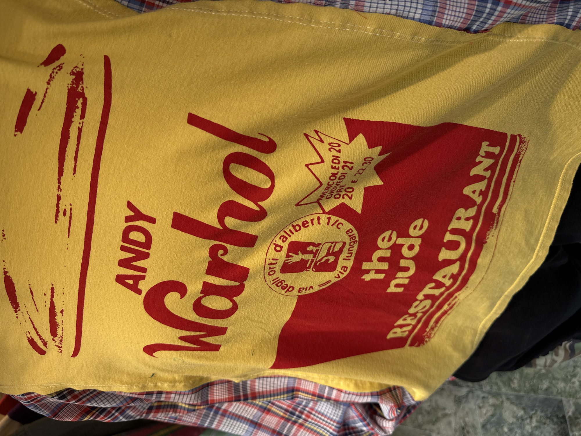 A yellow t-shirt with a distressed red print is prominently displayed, partially covering a plaid shirt with red, blue, and white squares. A dark-colored fabric is visible at the bottom right. The t-shirt's print features large, stylized text "ANDY Warhol" on the left side, reminiscent of pop art graphics. To the right, within a red rectangular design with a yellow border, there's further text and a circular emblem.
The text on the t-shirt includes:
- "ANDY Warhol"
- Inside a starburst shape: "MERCOLEDI 20", "GIOVEDI 21", "ORE 20 E 22.30" (indicating Wednesday 8 PM, Thursday 9 PM, and times 8 PM and 10:30 PM).
- Within a circular emblem, partially obscured but readable: "orti d'alibert 1/c", "via degli", and "via lungara". This points to an Italian location, likely Rome, given the street names Orti d'Alibert and Via della Lungara.
- Below the emblem: "the nude RESTAURANT".
The image appears to be an indoor shot of folded or stacked clothing. The t-shirt's fabric texture is visible, suggesting a worn or casual garment. There is no specific activity depicted. The lighting is even, and no time of day or specific weather is indicated.