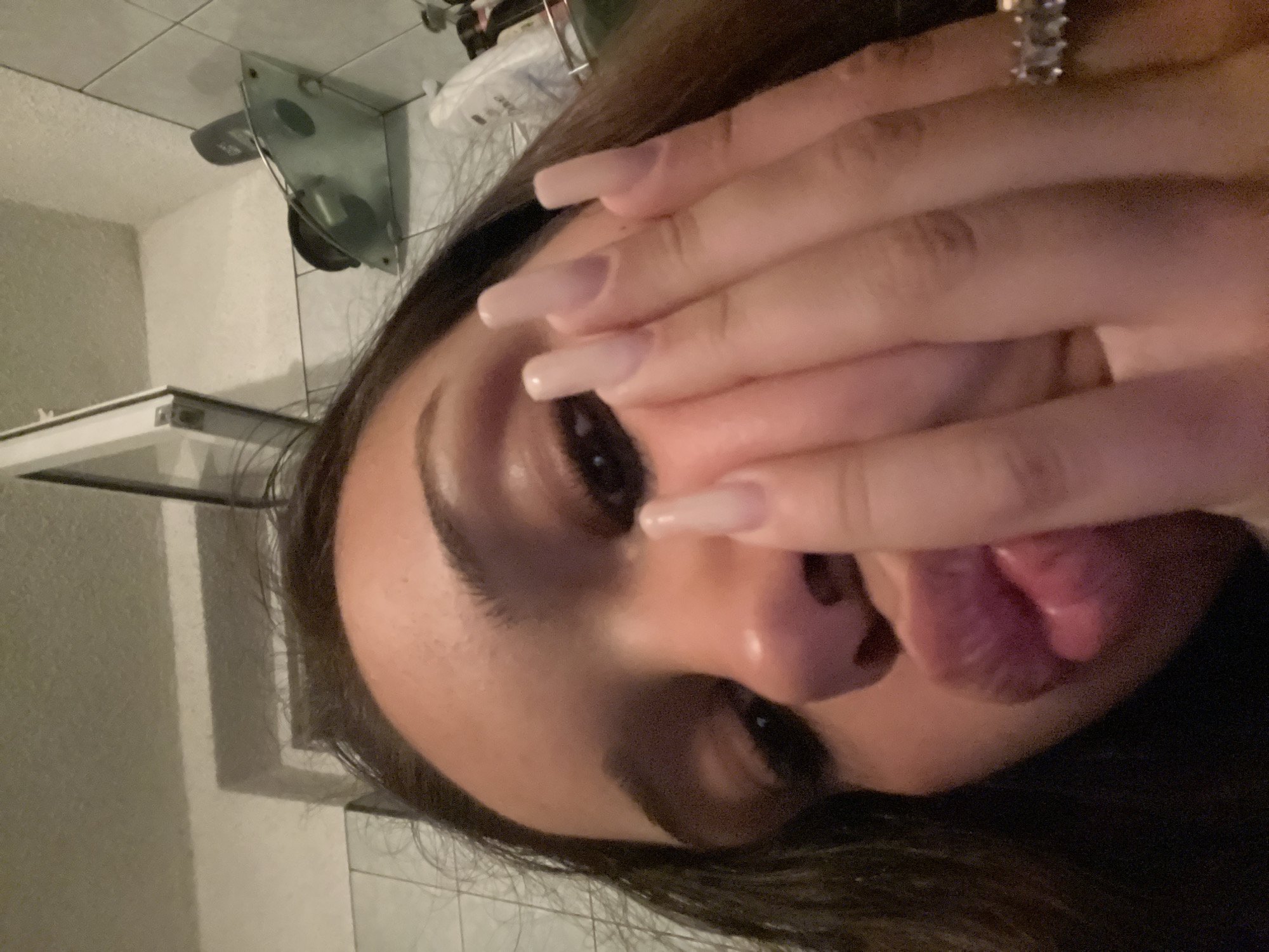 An upward-angled, close-up photograph depicts a young female subject indoors, with her lower face partially obscured by her right hand. The subject has long, dark hair, defined eyebrows, and visible eye makeup. Her lips are pursed in a pouting expression. Her right hand displays long, light-colored, artificial nails and a ring featuring multiple rectangular stones on the ring finger. The background consists of white tiled walls, a metallic-framed partition (possibly a shower door or mirror frame), and a glass shelf holding bottles, one of which bears a "Dove" brand label. The lighting is subdued and artificial. The specific city and region are unknown.