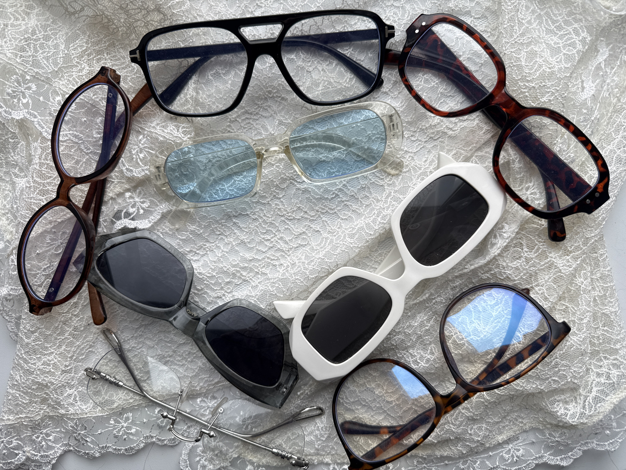 The image shows an arrangement of seven pairs of eyeglasses and sunglasses placed on a white lace fabric. The eyeglasses and sunglasses are of various styles, including black-framed rectangular glasses, tortoiseshell-patterned round glasses, blue-tinted glasses, gray-framed sunglasses, white-framed sunglasses, rimless glasses, and brown-framed round glasses. The scene appears to be staged, with the eyewear positioned aesthetically on the lace fabric. There is no indication of a location in Izhevsk, Russia.