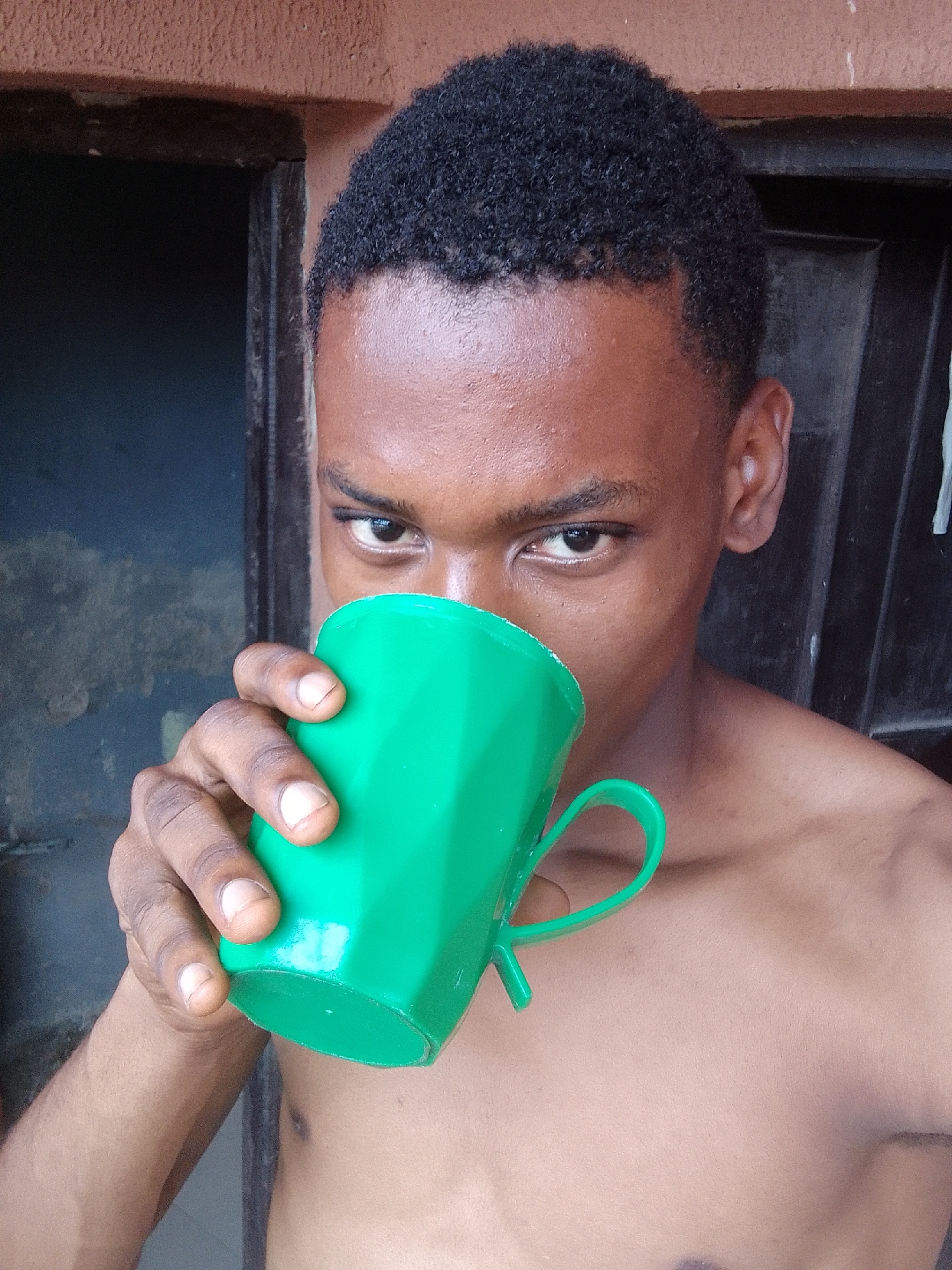 A shirtless young man is drinking from a green mug. The man has short, dark, curly hair and brown skin. The mug obscures the lower half of his face. The background is a doorway, with a dark interior. The setting is presumed to be within a building in the city of Unknown, Unknown.