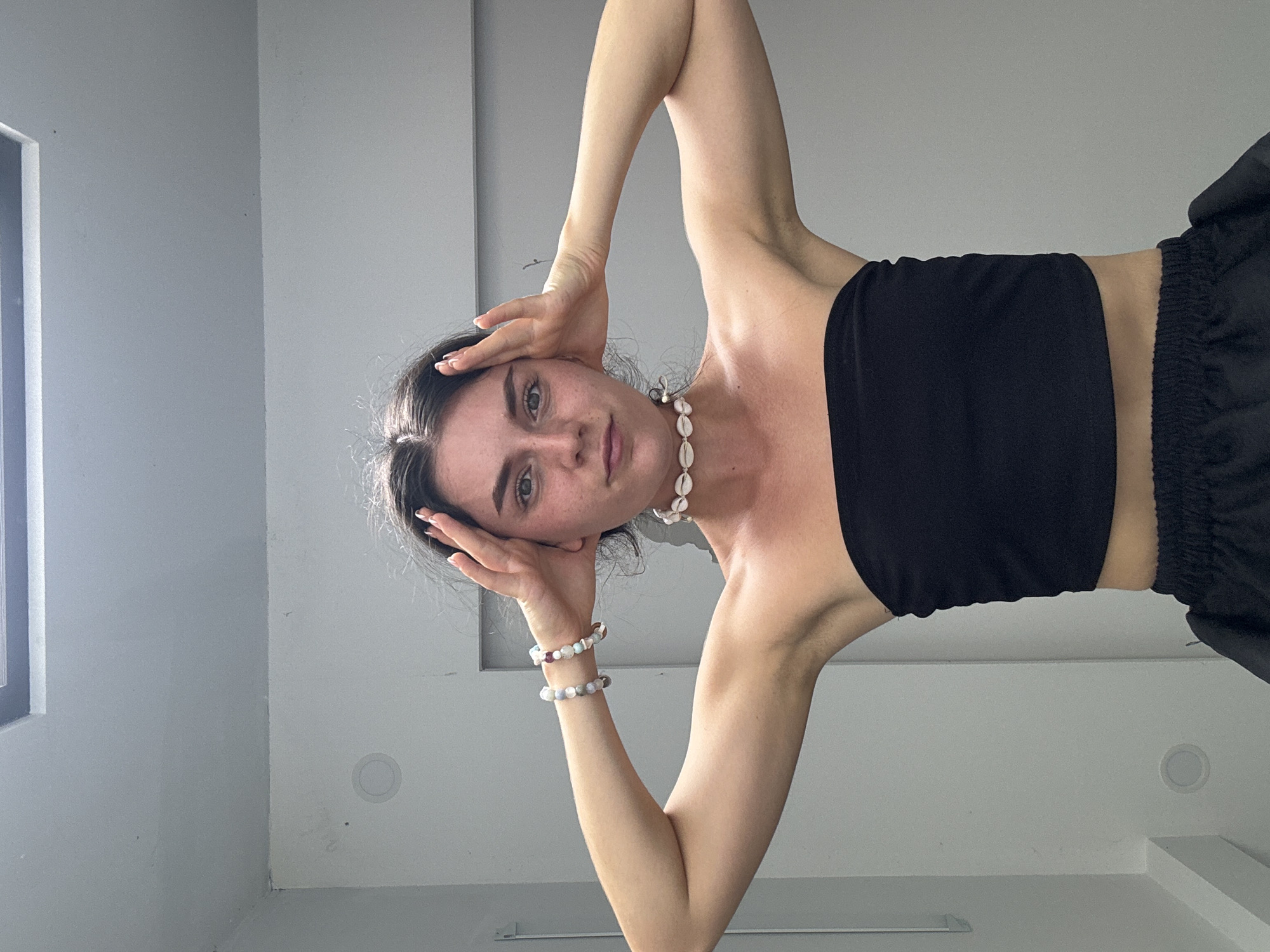 A woman is shown from a low-angle perspective indoors. She has light skin, dark hair, and is wearing a black tube top and black shorts. Her hands are positioned on either side of her head, and she is wearing a shell necklace and two bracelets. The background consists of white walls and a window. The location is unspecified, but the prompt mentions Mũi Né, Vietnam, without specifying that this image was taken there.