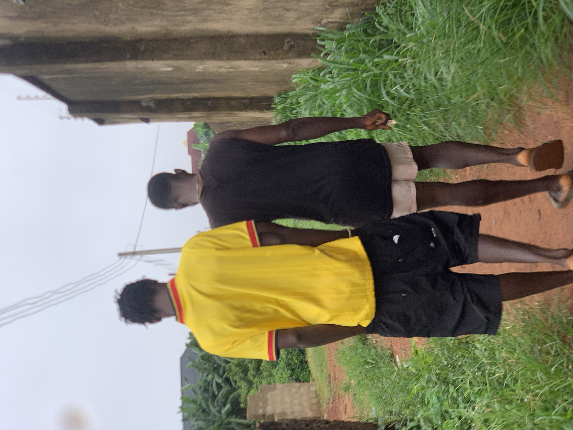 Two individuals, seen from behind, walk along a dirt path in what appears to be Ukwu-Abwa, Nigeria. The person on the left wears a bright yellow t-shirt with red and black trim on the collar and sleeves, and black shorts. The person on the right is wearing a black sleeveless t-shirt and light-colored shorts. The environment is characterized by lush green vegetation lining both sides of the path, and a textured, aged wall runs along the right side of the frame. Overhead, power lines are visible against an overcast sky. The ground is a reddish-brown dirt.