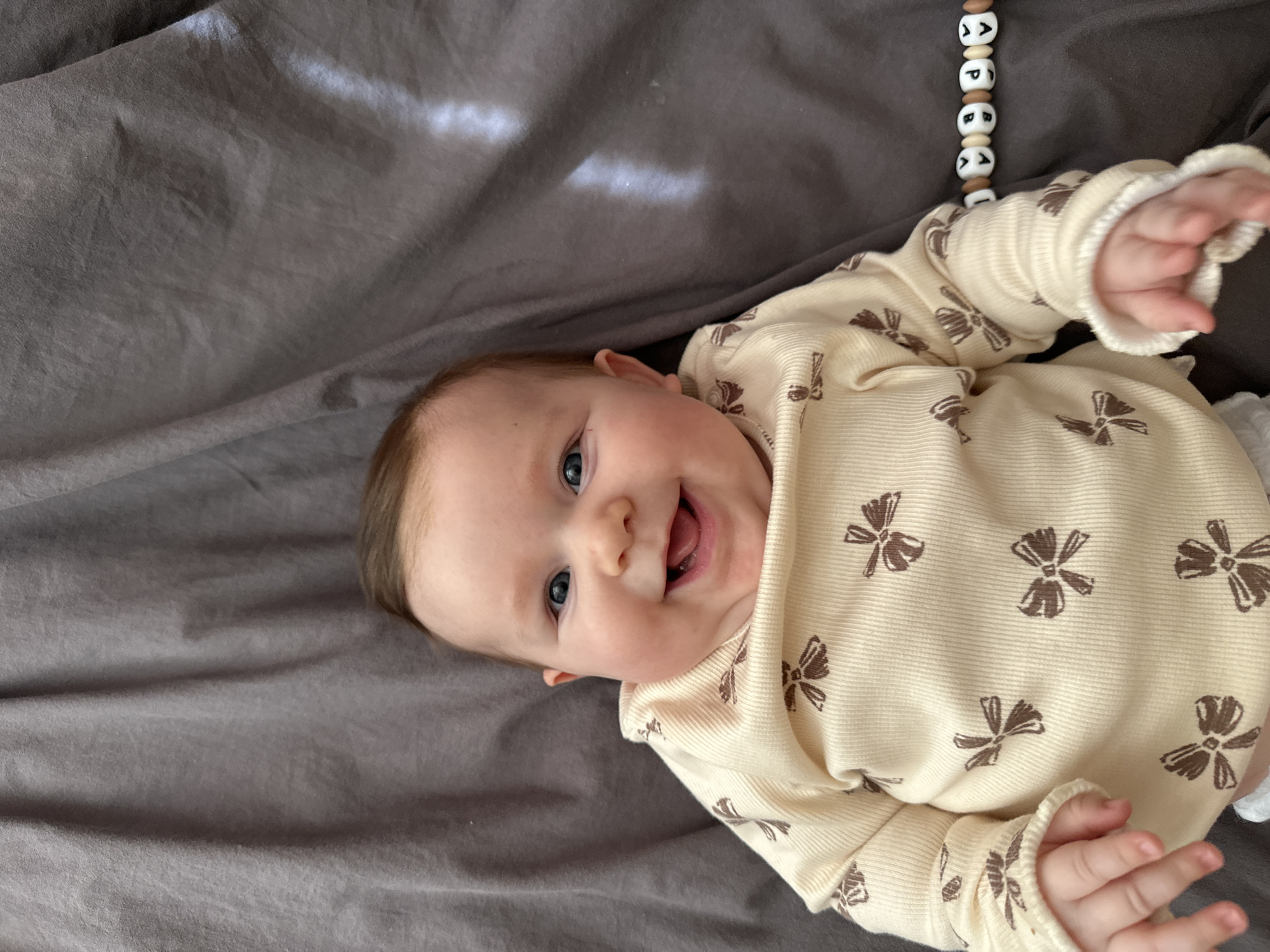 A baby lies on a grey sheet, wearing a light-yellow onesie with brown bow patterns. The baby is smiling, revealing two lower teeth. Above the baby is a string of wooden beads. The location is indiscernible.