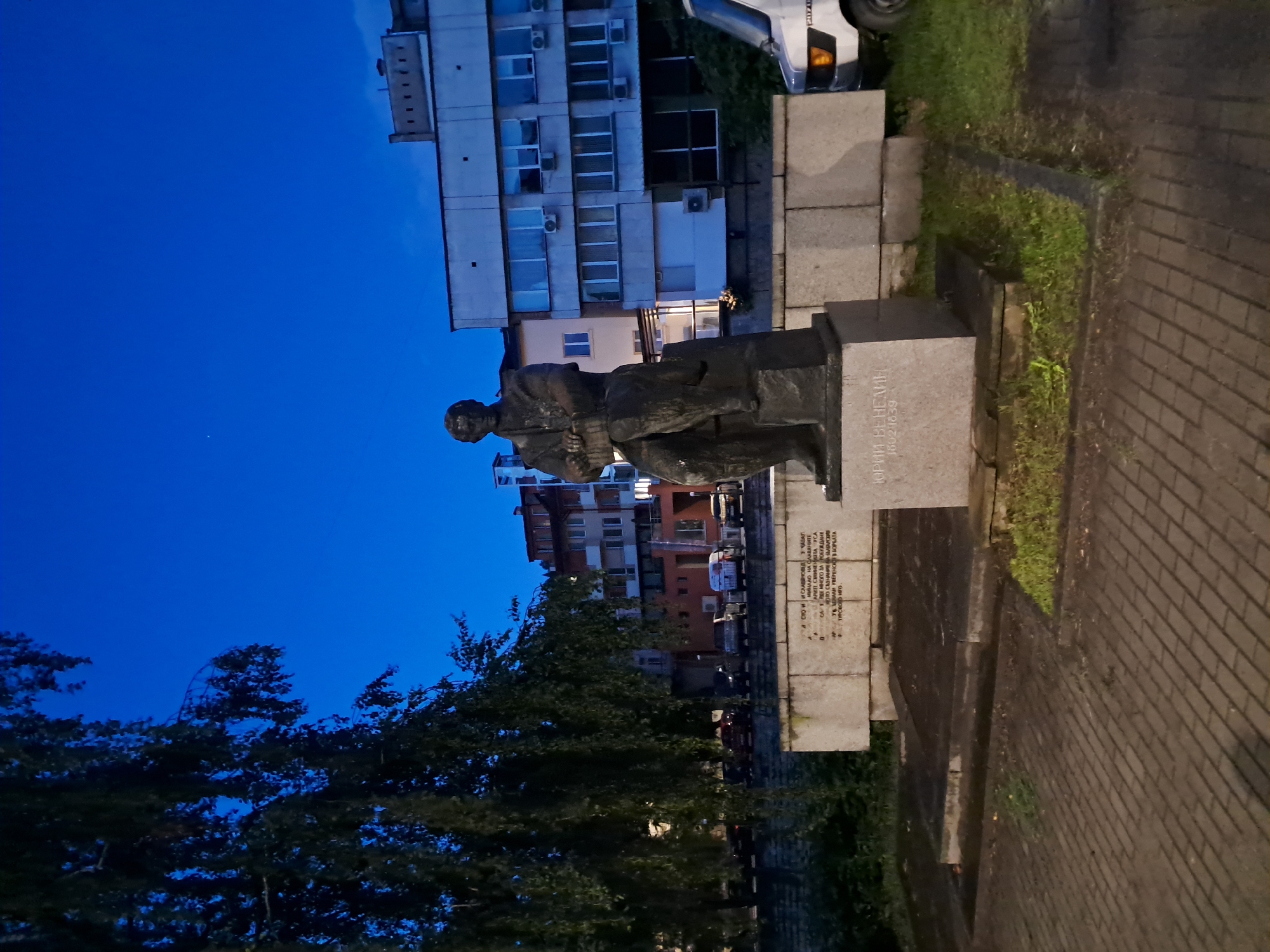 The image captures a bronze statue of a man, likely a historical figure, standing on a stone pedestal in an urban setting at dusk or night. The statue depicts a person dressed in formal or traditional attire, holding what appears to be a book or scroll in their left arm. Behind the statue, a multi-story building with numerous windows and air conditioning units is visible, illuminated by interior lights. A portion of a white or silver car is parked to the right of the building.
The foreground features paved ground, resembling brickwork, and patches of grass. Large, dark trees line the left side of the frame, silhouetted against the deep blue sky. The sky is clear, indicating fair weather. The overall lighting suggests it is late evening or early night.
Visible text on the pedestal, translated from Cyrillic, includes the name "ЮРИЙ ВЕНЕЛИН" (Yuriy Venelin) and the numbers "16021839", likely representing his birth and death years. Another inscription, partially visible, contains phrases like "СЛАВЯНОВЕД" (Slavist), "МИНАЛО НА СЛАВЯНИТЕ" (Past of the Slavs), "ПРОБУЖДАНЕ" (awakening), and "ТУРСКОТО ИГО" (Turkish Yoke), suggesting the monument honors Venelin's contribution to Slavic studies and the national revival from Ottoman rule.
