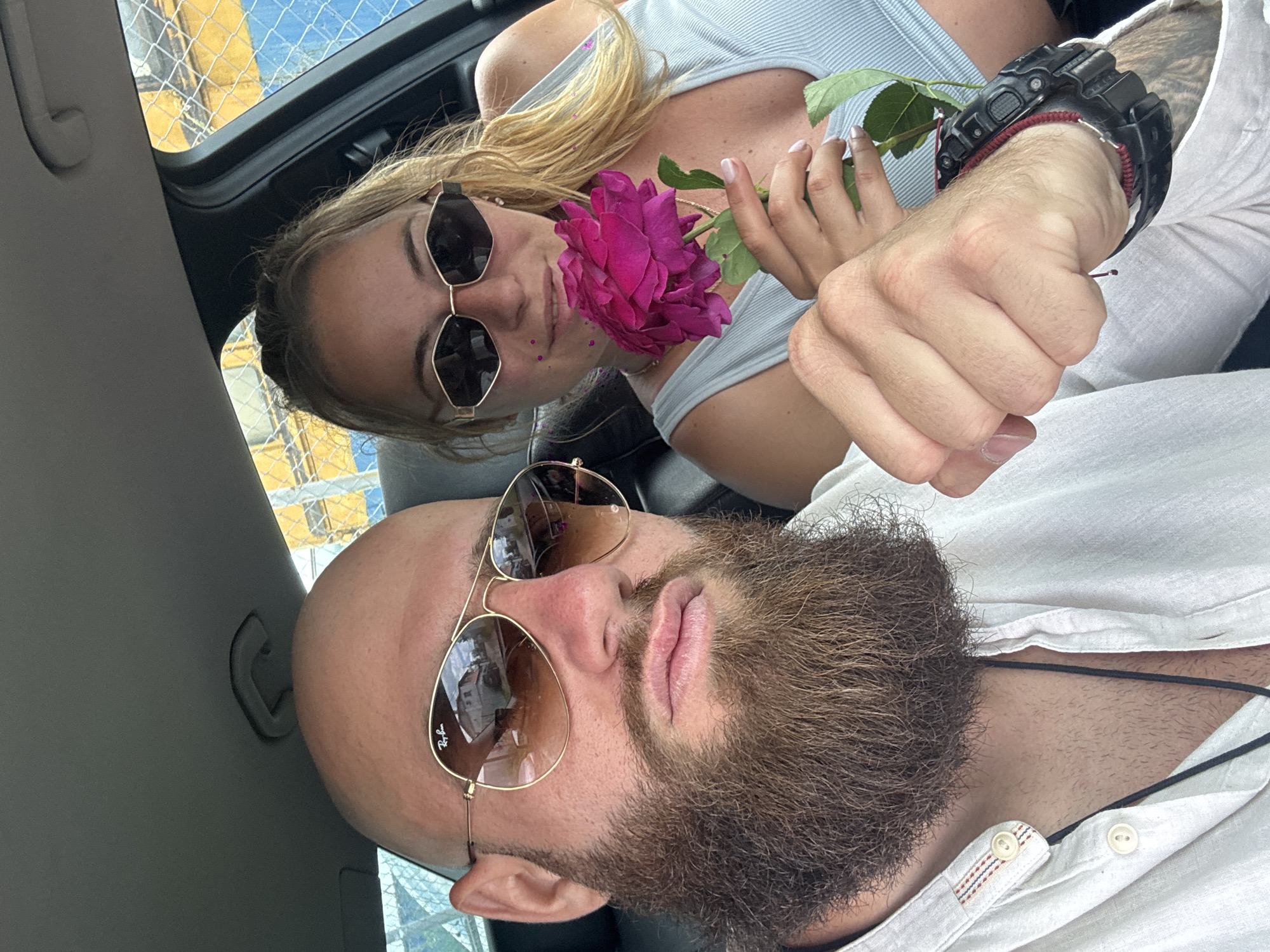 The image shows two individuals in the interior of a vehicle. A woman with blonde hair and sunglasses is holding a purple rose. A man with a beard and sunglasses is next to her. Both are wearing light-colored clothing. The background visible through the vehicle window includes what appears to be a metal fence and a glimpse of buildings in the distance. The location appears to be Unknown, Unknown.