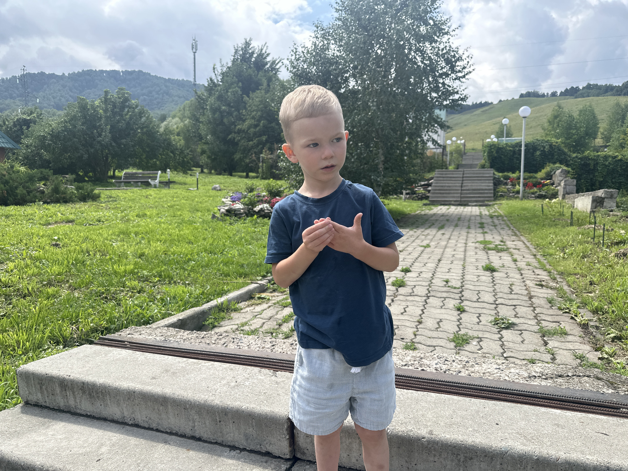A young boy with blonde hair and blue eyes stands facing right, wearing a dark blue t-shirt and light blue striped shorts. He is holding his hands clasped in front of him. He is positioned on a concrete structure in what appears to be a park or recreational area. The background features a paved walkway with a zig-zag pattern, surrounded by green grass and trees. In the distance, there is a hillside with more trees and a cloudy sky. The location is identified as Danilovka (Altai), Russia.