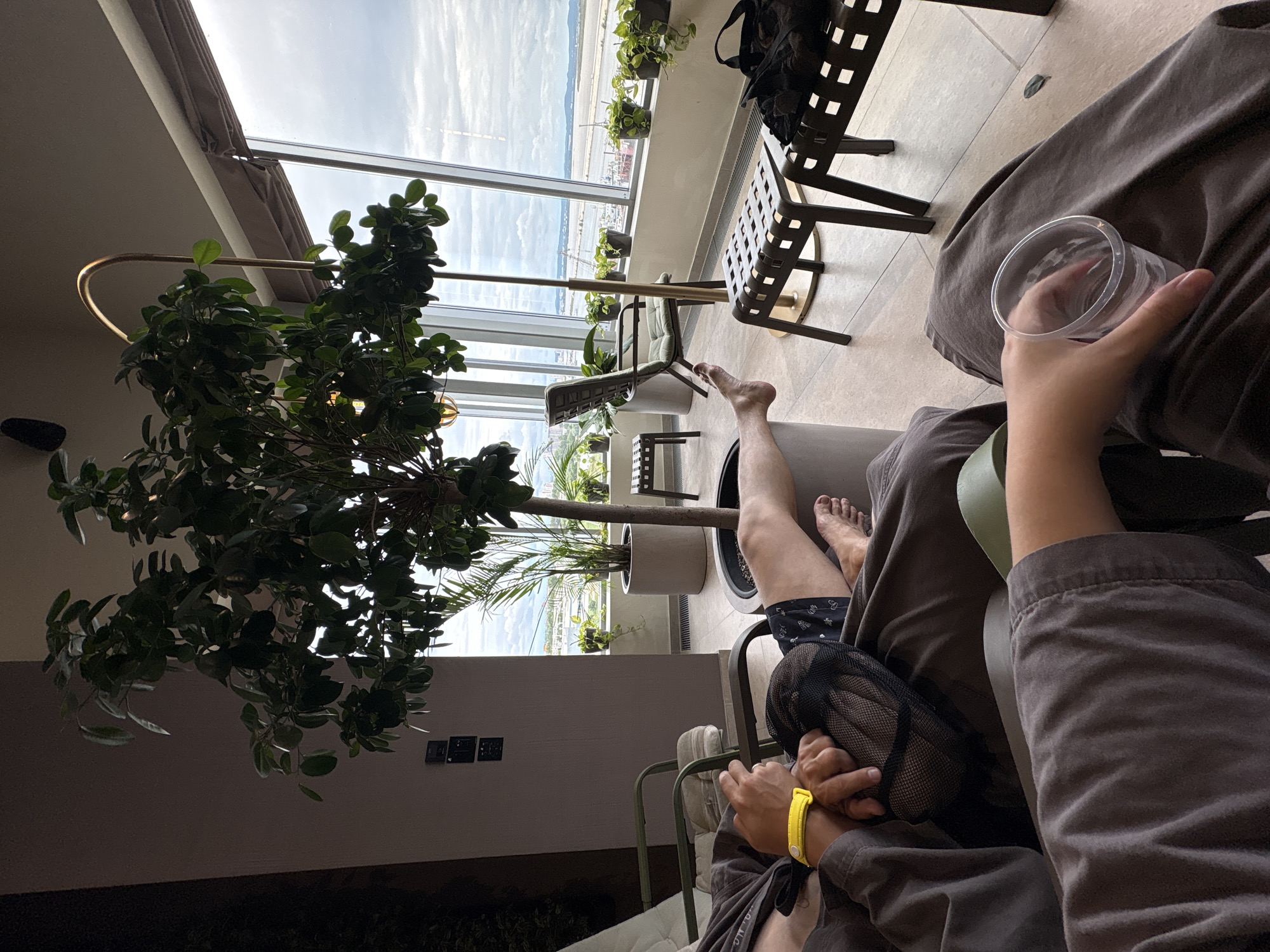 The image depicts a lounge area with large windows offering a view of a river or bay and a cloudy sky, consistent with the geography of Khabarovsk, Russia. In the foreground, two individuals are seated in lounge chairs. One person, wearing a grey robe and a yellow wristband, is visible from the waist up, holding a black mesh bag. Their legs are extended. The second person's arm and hand are in the lower right of the frame, holding a clear plastic cup.
The room is furnished with modern seating, including a green-cushioned chair and several black lattice-style ottomans. A tall potted plant with lush green leaves dominates the central part of the scene, positioned in front of the windows. Other smaller potted plants are arranged along the windowsill. The flooring appears to be light-colored tile. Electrical outlets are visible on the wall. The overall atmosphere suggests a relaxation or waiting area, possibly at a spa, airport lounge, or similar facility.