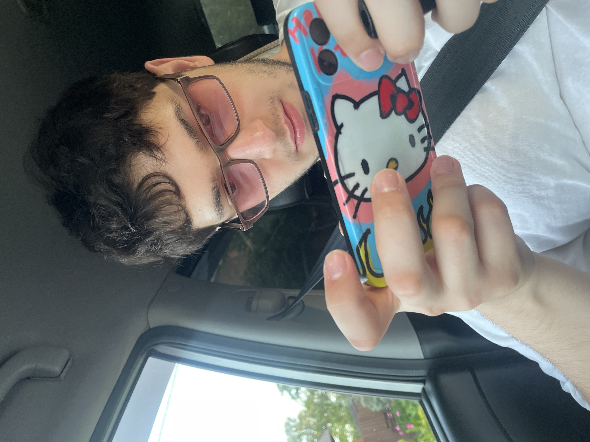 A young man with dark, curly hair and pink-tinted glasses is holding a smartphone with a Hello Kitty case in a car. He is wearing a white t-shirt. The car's interior is visible, including a window showing greenery and a house outside, suggesting a suburban or rural setting, possibly in Votkinsk, Russia. The man's hands are positioned as if he is using the phone. A seatbelt is also visible.