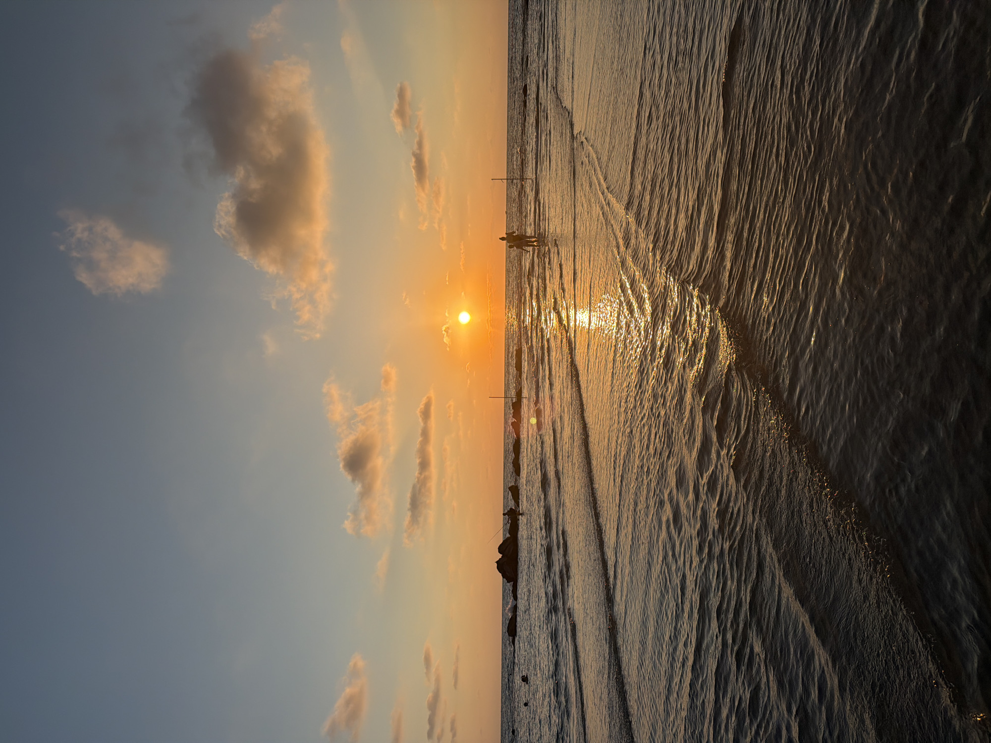 A sunset over the Mediterranean Sea in Bat Yam, Israel. The sun is a bright orange orb low on the horizon, casting a golden reflection on the water. The sky is a gradient of blue and orange, with a few wispy clouds catching the sunlight. In the foreground, gentle waves lap at the shore, creating a rippling texture on the water. In the middle ground, two people are walking in the shallow water, silhouetted against the setting sun. Further out, a line of rocks breaks the water's surface, and a lone figure can be seen fishing near them. The scene is tranquil and picturesque, capturing the beauty of a Mediterranean sunset.