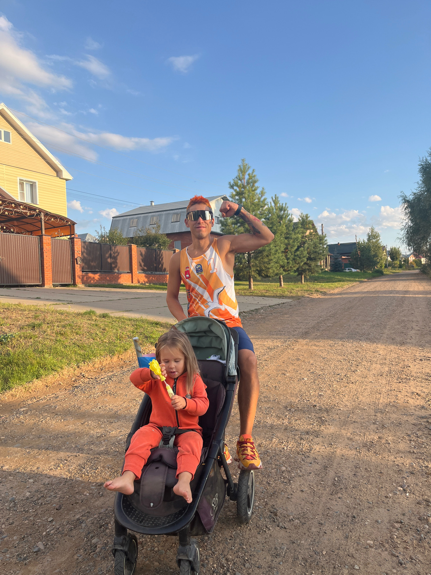 A young, fair-skinned man with bright orange hair and sunglasses is posing while pushing a stroller with a young child in it. The man is wearing a sleeveless orange and white athletic tank top and blue shorts. His left arm is flexed, showing a tattoo on his bicep. The child, a young girl with blonde hair, is sitting in the stroller and holding a yellow toy. She is wearing an orange jumpsuit and is barefoot. They are on a gravel road in a rural or semi-rural area, possibly on the outskirts of the city of Chaikovsky, Russia, given the presence of houses with pitched roofs and fences, as well as mature trees and a clear blue sky with scattered clouds. The sun appears to be setting or rising, casting long shadows. The scene suggests a casual outing or a walk.