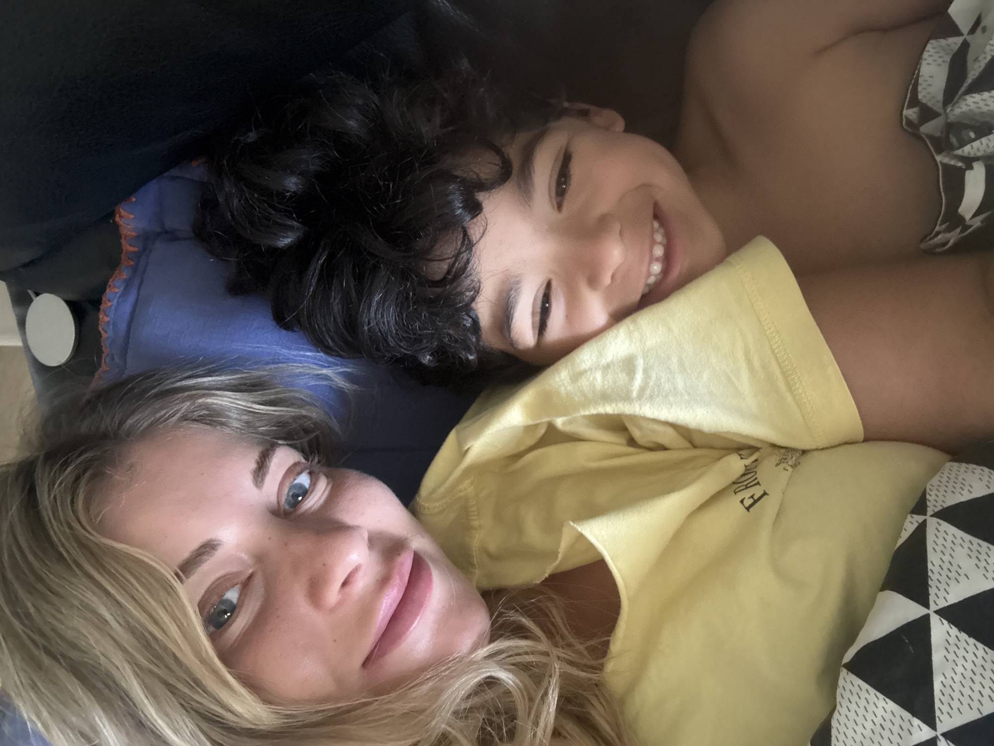 A mother and child are lying next to each other, possibly on a bed or couch. The mother, who has blonde hair and blue eyes, is wearing a yellow t-shirt with the words "FROM" and "RO" visible. The child, who has dark curly hair, is smiling and has a blanket with a black and white geometric pattern around them. A blue pillow is visible under the child's head, and the background includes a dark-colored couch. The photograph was probably taken in Rousset, France.