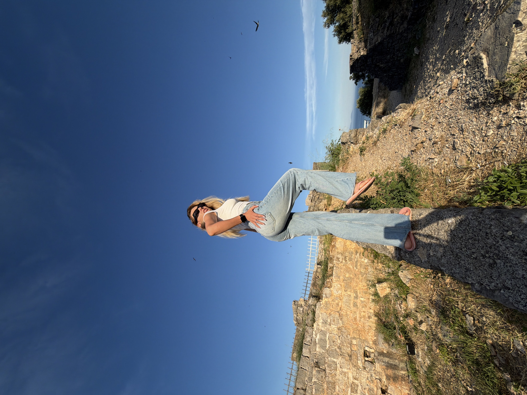 In Budva, Montenegro, a young woman with blonde hair stands on a weathered stone wall overlooking the Adriatic Sea. She is wearing a white tank top and light-wash flared jeans, her right hand resting on her hip. The woman is also wearing sunglasses and pink sandals. The sky is a vibrant blue with a few scattered birds flying. The background features ancient stone ruins and lush green vegetation, suggesting a historical site. The lighting indicates it is late afternoon or early evening, with the sun casting long shadows.