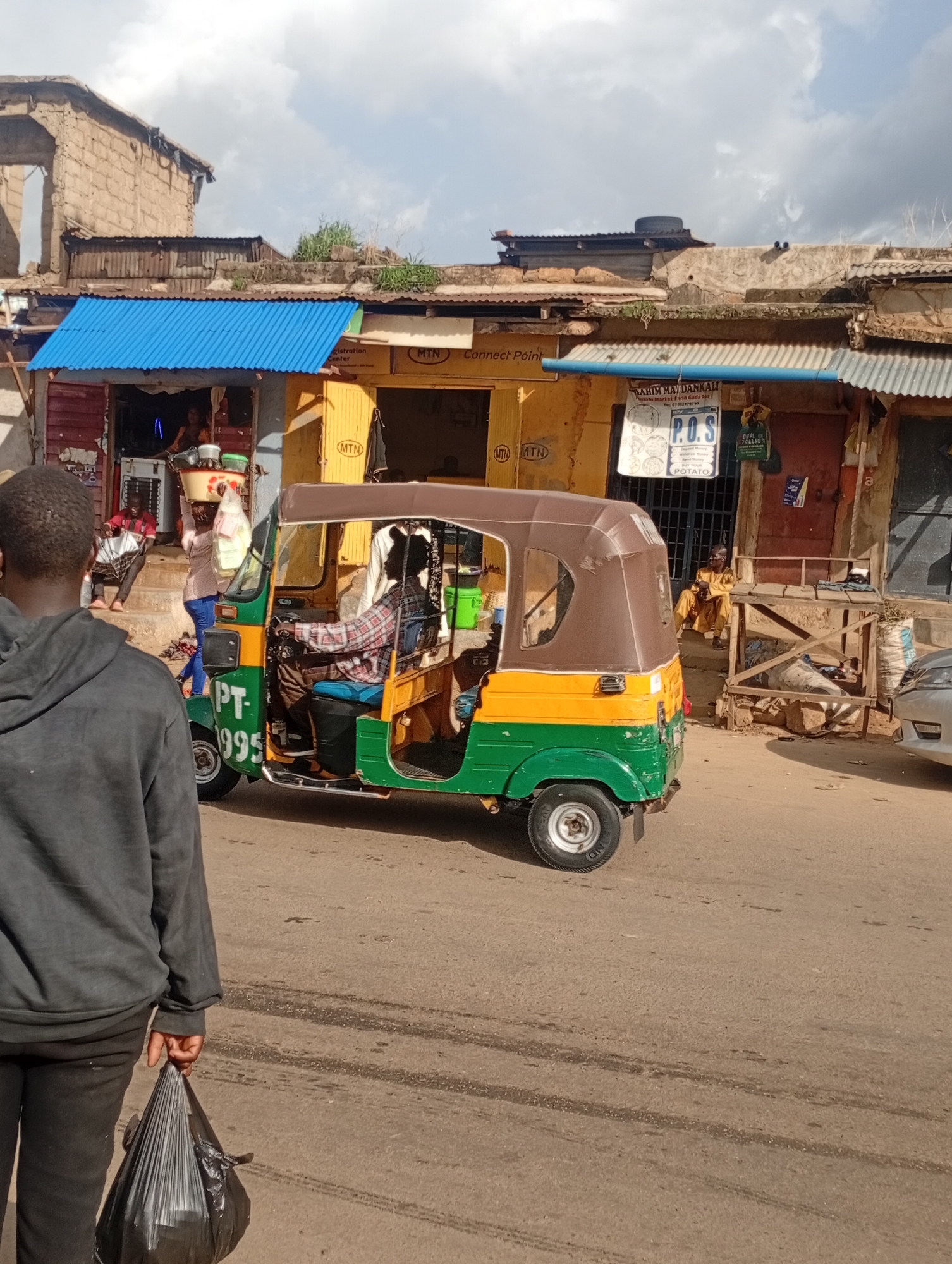 The image shows a street scene in Rafin Par, Nigeria, with a tuk-tuk vehicle as the central focus. A person, viewed from behind, walks away from the camera holding a black plastic bag. The tuk-tuk, a three-wheeled auto-rickshaw painted green and yellow, is in motion or parked on the right side of the street. The driver, wearing a plaid shirt, is visible behind the steering wheel. Behind the tuk-tuk, there are several small shops or stalls with corrugated metal roofs and various signage. One stall has a blue awning. A woman carrying a bowl of items on her head walks away from a shop. Another person in a yellow garment is seated in front of a stall. The environment appears to be a busy marketplace with buildings constructed from concrete blocks and corrugated metal. The sky is partly cloudy.