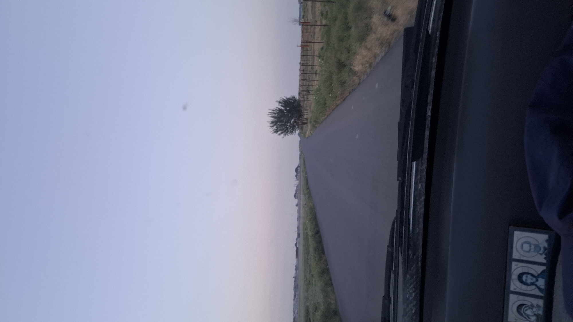 The image depicts a view from inside a vehicle, looking out onto a rural road in Chubarsu, Kazakhstan. The scene is captured at dusk or dawn, with the sky transitioning through pale blues and pinks. A paved road stretches into the distance, flanked by grassy fields and a fence line. A lone tree stands prominently on the left side of the road. The dashboard of the vehicle is visible in the foreground on the right, with a dark interior. On the dashboard, a small framed item with three portraits is visible, featuring stylized depictions of individuals. The overall impression is one of a quiet, perhaps solitary journey through a peaceful landscape. The time of day suggests either the beginning or end of daylight hours, creating a soft, diffused light across the scene. No specific activities or emotions are explicitly conveyed, beyond the implied movement of travel.