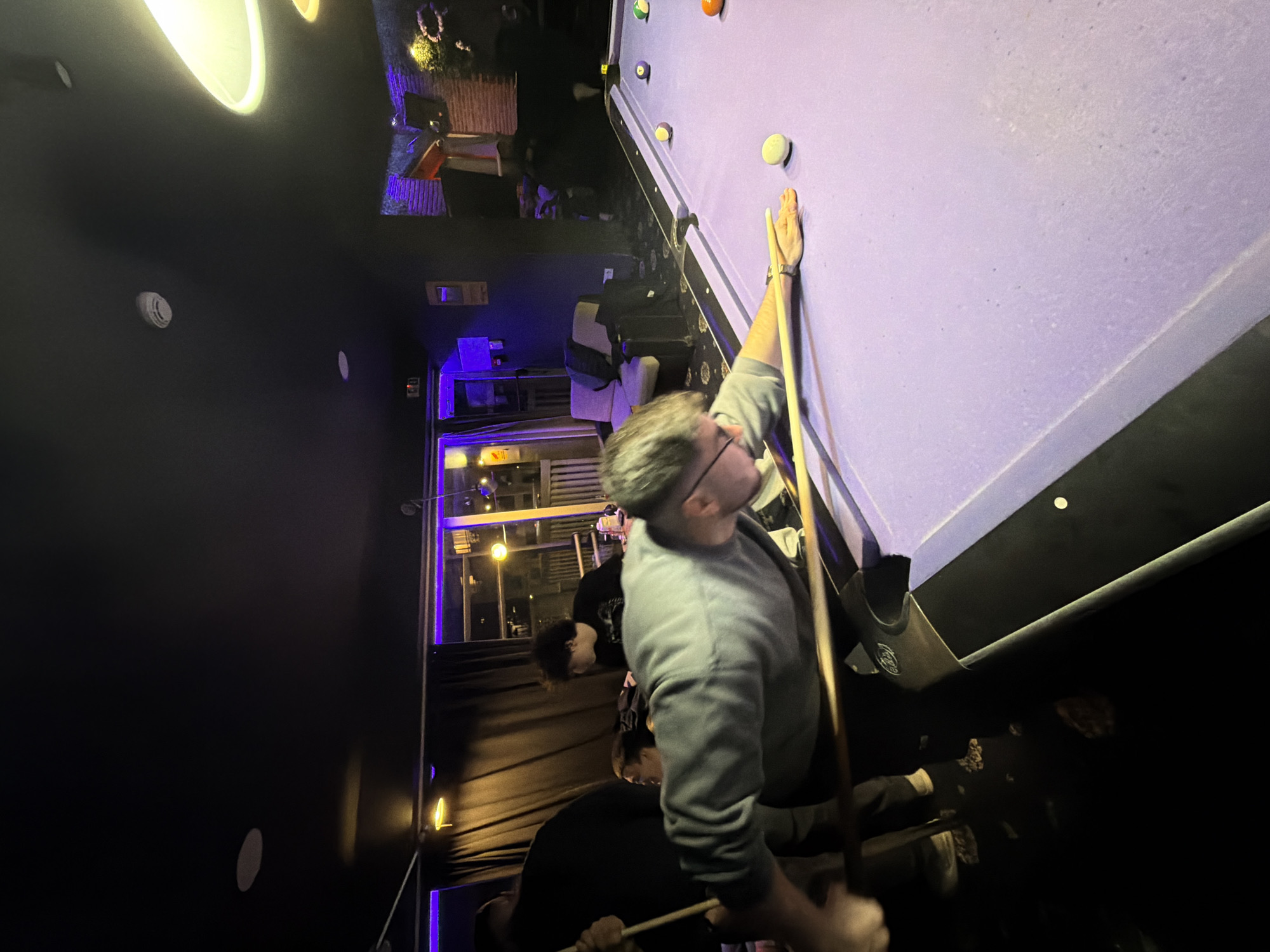 A group of people are playing pool in a dark indoor location. One man with glasses and gray hair, wearing a gray sweater, is leaning over the pool table, cue in hand, preparing to strike a white ball. The pool table is covered in lavender felt and has several other balls on it. Other people are standing around, observing the game. There are black walls with a few visible lighting fixtures. Through a doorway in the background, there are glimpses of an outdoor area with string lights and what appears to be the exterior of a building, suggesting a bar or entertainment venue in the city of Plovdiv, Bulgaria.