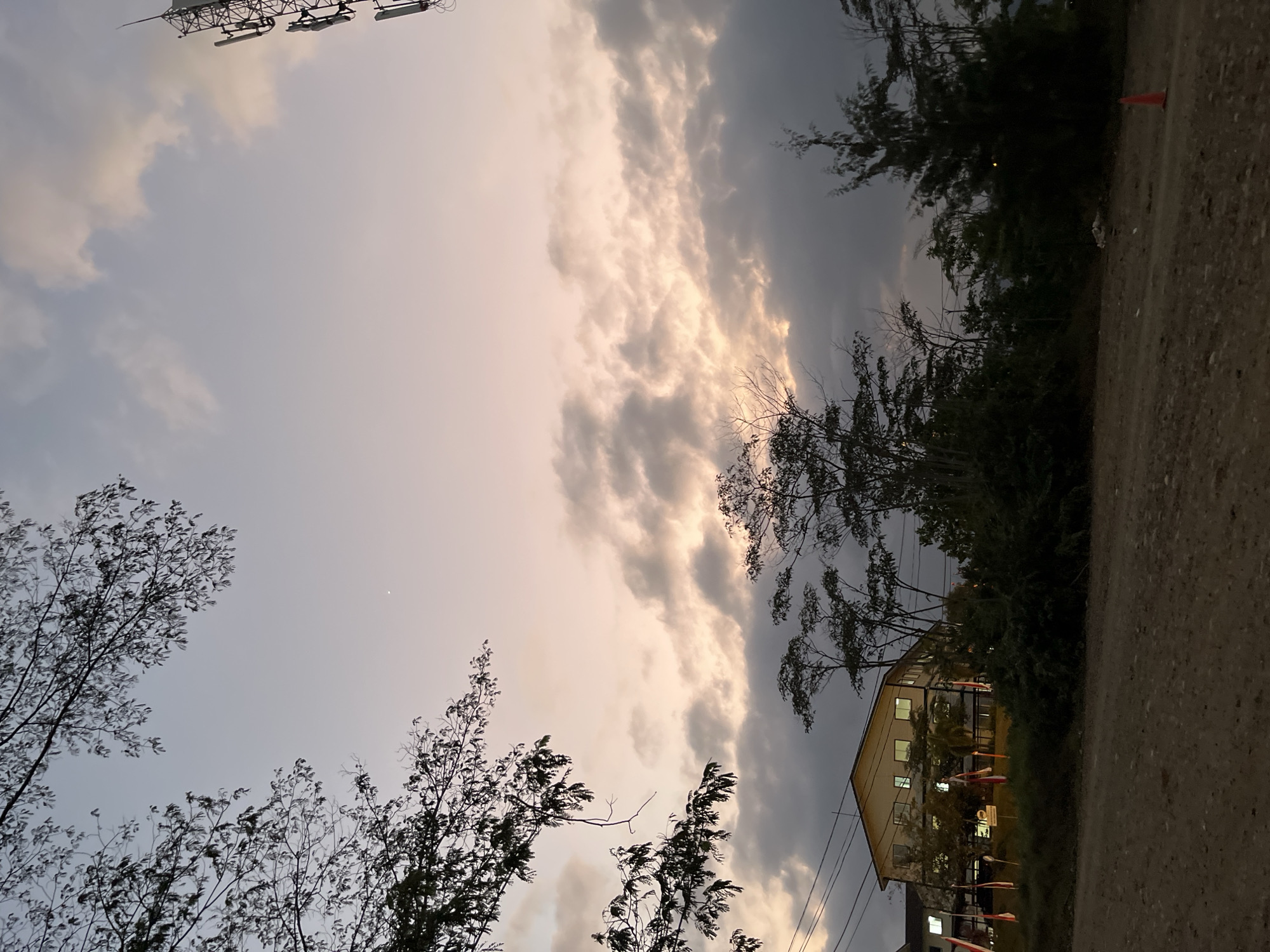 The image shows a twilight scene in Lalampu, Indonesia. In the foreground, a dirt road curves towards the right, with an orange traffic cone visible near the lower right corner. Beyond the road, there is a dense area of trees and bushes. A multi-story building with cream-colored walls and illuminated windows stands on a slightly elevated embankment, flanked by trees and several Indonesian flags displayed on poles. Power lines are visible running horizontally above the building and trees. In the background, the sky is filled with a mix of soft pastel clouds tinged with the warm hues of sunset, and darker, more ominous clouds. To the right of the frame, a tall telecommunications tower with multiple antennas is prominently positioned against the sky. The overall atmosphere suggests the transition from day to night in a semi-urban or suburban environment.