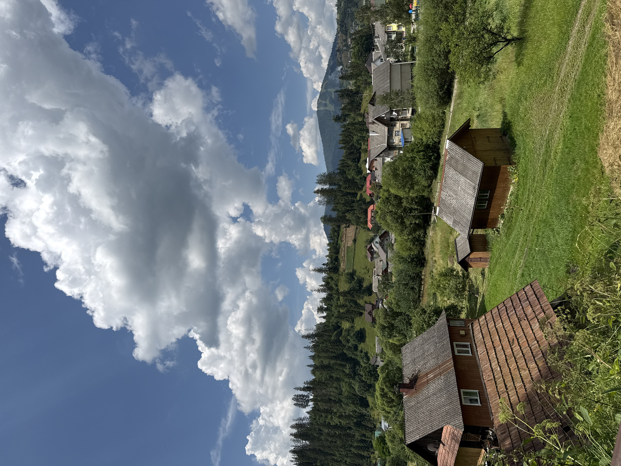 A rural village scene in Slavsko, Ukraine, is depicted under a bright blue sky dotted with voluminous white clouds. The foreground features the weathered, terracotta-tiled roof of a traditional wooden house, partially obscured by lush green vegetation. To the right, a smaller, rustic wooden shed with a grey, corrugated roof sits on a gently sloping, verdant lawn.
Further into the frame, a cluster of residential buildings with varied architectural styles and roof types forms the heart of the village. These houses are nestled amongst mature trees and greenery. Beyond the village, a dense forest blankets a rolling hillside, creating a striking contrast with the cleared land and buildings. In the background, a majestic mountain range rises, its slopes covered in evergreen trees, with a visible ski lift or trail indicating the area's recreational use. The overall impression is one of a serene, picturesque mountain community.