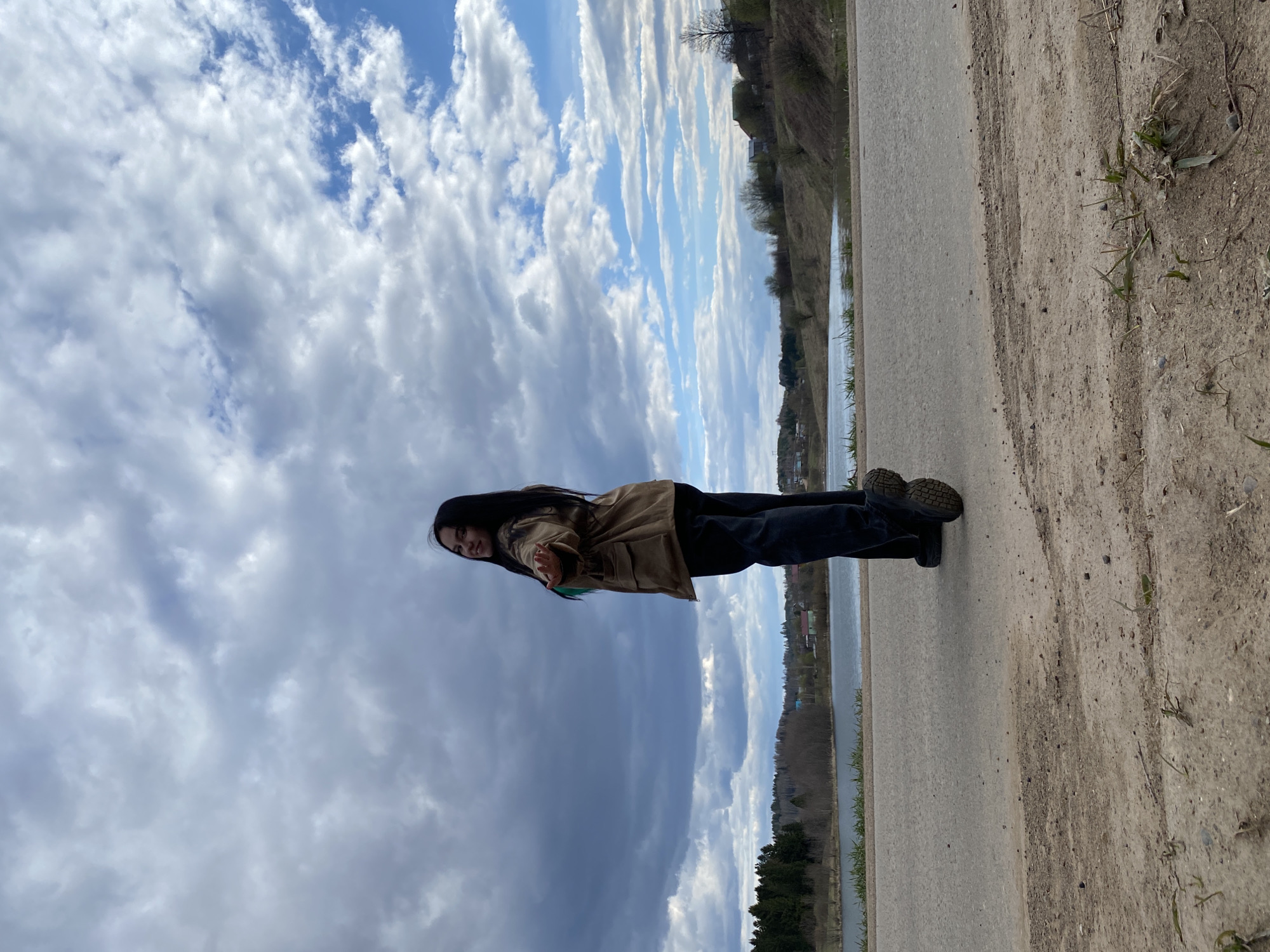 The image, correctly oriented, depicts a young woman standing outdoors in what appears to be a rural or semi-rural setting in Фоки, Russia. She has long dark hair and is wearing a light brown or khaki jacket, dark trousers, and sturdy dark boots. She is positioned on a dirt path, standing next to an asphalt road that runs parallel to a body of water, likely a river or lake. The woman is looking towards the camera with a slight smile and has her right hand extended slightly forward, as if in a gesture or pose.
The sky above is prominent, featuring a dynamic mix of bright white and grey clouds, with patches of clear blue sky visible, suggesting a day with variable weather. The time of day appears to be daytime, likely late morning or early afternoon, given the natural lighting.
Across the body of water, the distant shoreline is visible, characterized by a line of trees and some scattered buildings, indicating a modest settlement further along the bank. The immediate foreground on the right consists of a rugged dirt and gravel path with sparse green grass and weeds. There is no visible text in the image. The scene conveys a calm, natural outdoor environment.