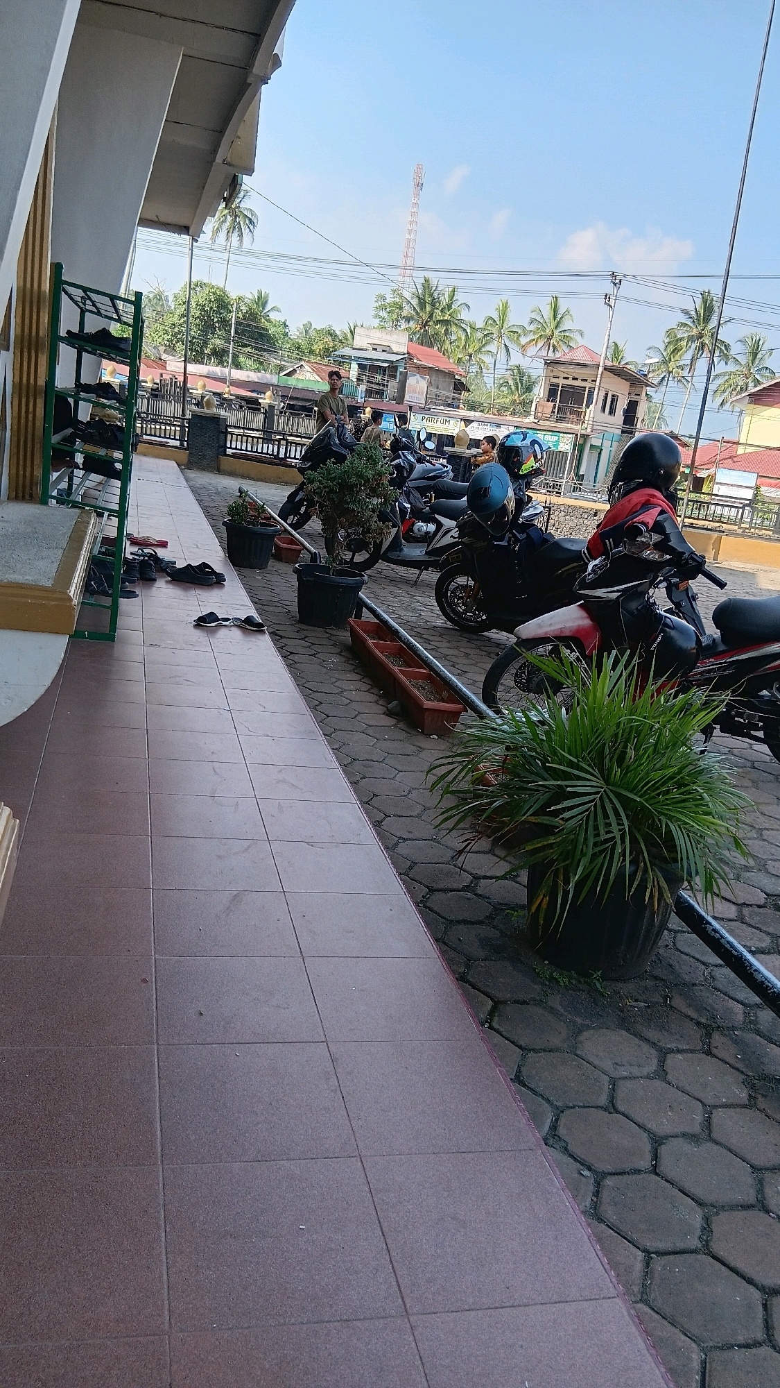 The image depicts an outdoor scene in Sungai Tarab, Indonesia, likely in front of a building. The foreground shows a tiled walkway with several pairs of footwear scattered on it, suggesting a place where shoes are removed. To the right of the walkway, a paved area with hexagonal stones is visible, hosting a row of parked motorcycles. Potted plants, one with lush green foliage, are placed between the motorcycles. In the background, more motorcycles are parked, some with helmets placed on them. A few individuals are present, one standing near a motorcycle, and another further in the background. The environment appears to be a commercial or public area, with buildings and palm trees visible. A cell tower stands prominently in the distance against a clear blue sky with a few wispy clouds. The scene suggests a typical day in a warm climate, with activity around parked vehicles.
