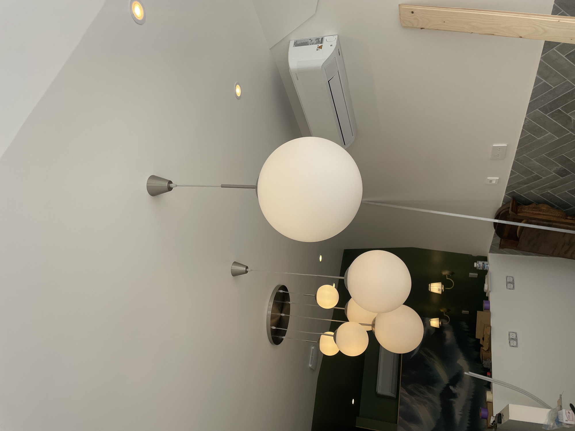 The image depicts an interior scene, likely a room under renovation or recently completed. A white ceiling dominates the upper portion of the frame. Several large, spherical pendant lights with opaque white globes hang from the ceiling, some in a cluster and one solitary light prominently in the foreground on the right. These lights have metal fixtures and cords. A silver-toned recessed light fixture is visible on the ceiling to the right of the foreground pendant light. An air conditioning unit is mounted on the wall to the right, below the ceiling.
To the left, the wall is painted a dark green and features a landscape mural with mountains and a body of water. Below this mural, on a white structure, are two wall-mounted sconces with cream-colored shades. Two electrical outlets are visible on the white structure. The lower section of the walls features a herringbone pattern of dark gray tiles.
In the lower right of the image, a vertical piece of light-colored lumber leans against the wall. The overall environment suggests a modern interior space with some ongoing or recent construction work, given the exposed electrical outlets and the lumber. There are no people visible in the image. The scene is well-lit, with the pendant lights casting a soft glow. No specific location in Meaux, France, can be determined from the image.