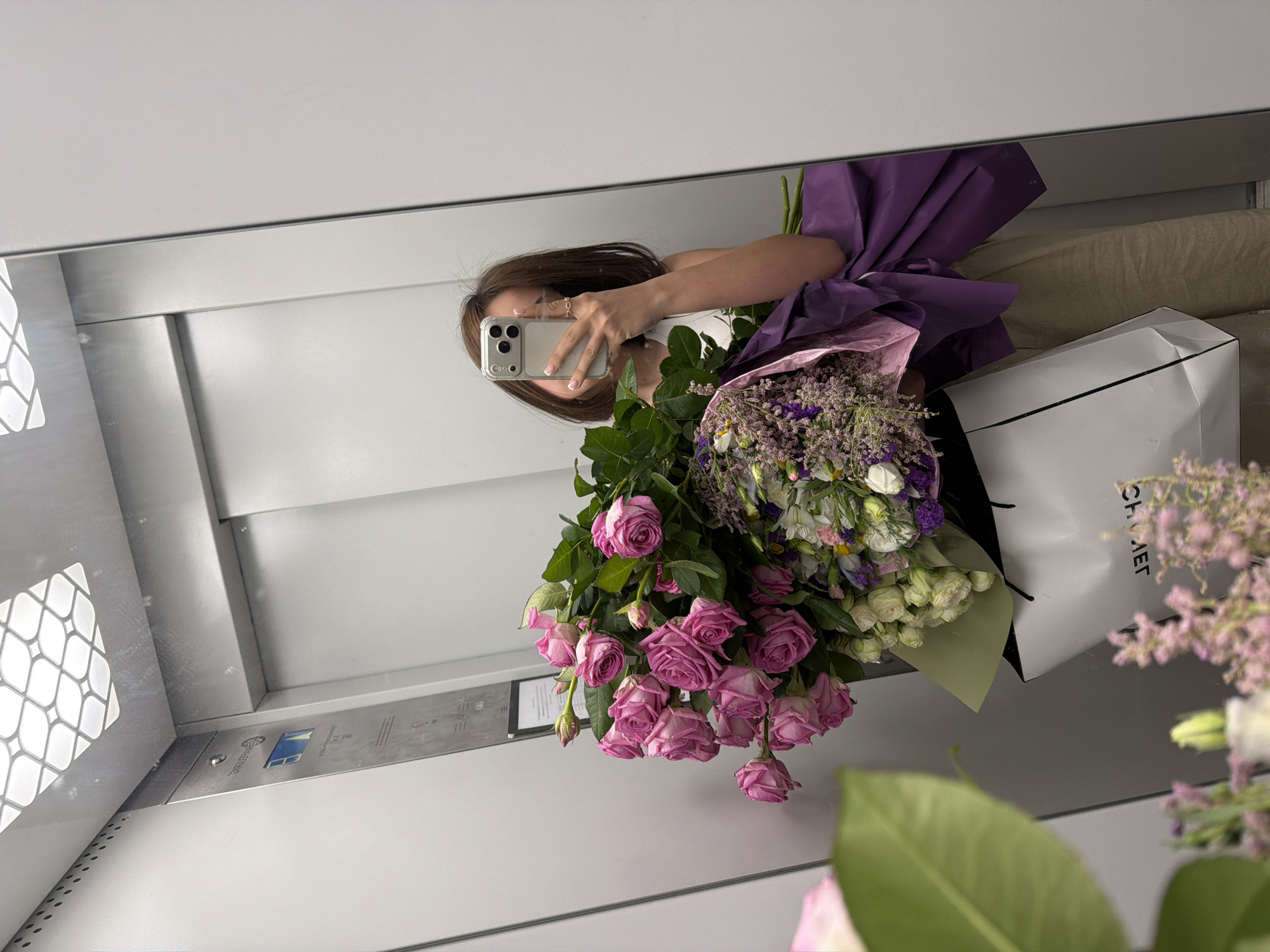 A mirror selfie captures a woman holding a bouquet of flowers and a shopping bag. The setting appears to be an elevator in Yerevan, Armenia, based on interior features and the possible urban context. The woman's face is partially obscured by her smartphone as she takes the photo. A variety of flowers, including roses and other blooms, are visible in the bouquet. The shopping bag, with the brand name "CHANEL," is present. The elevator has light-colored walls and metallic accents.