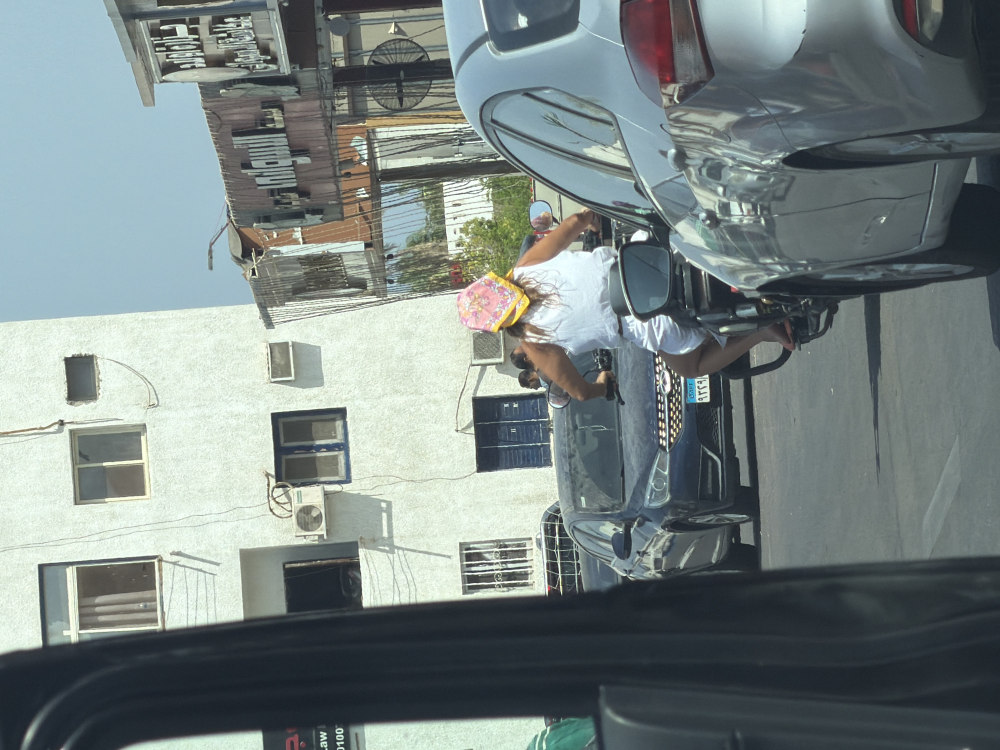 The image captures a street scene in Sharm El Sheikh, Egypt, on a sunny day. The foreground is dominated by a dark vehicle, likely a car, seen from the interior looking out. To the right, a silver car is partially visible, showing its rear quarter panel and taillight. In the middle ground, a person is riding a motorcycle, viewed from behind. They are wearing a white tank top and white shorts, with a bright pink and yellow bandana covering their head. The motorcycle is a dark color, and a side mirror reflects the scene behind. The person's feet are visible, wearing sandals.
Behind the motorcycle, a dark blue sedan is stopped in traffic. Its license plate, with Arabic characters and numbers, is partially visible. Further in the background, there is a white building with several windows, some with visible curtains or blinds, and an air conditioning unit mounted on the exterior. To the right of the white building, there are other structures with signs displaying Arabic text. The sky above is clear and blue. The overall impression is a typical urban street scene in Egypt, likely during daytime, with traffic and pedestrian activity.