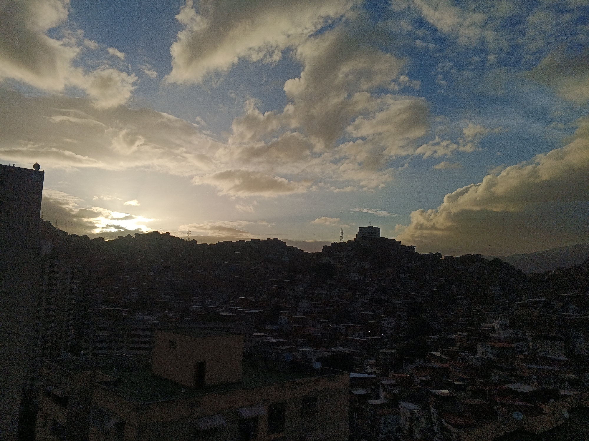 The image captures a vast urban landscape at sunset, showcasing a densely populated hillside settlement in Caracas, Venezuela. The sky dominates the upper half of the frame, featuring a blend of deep blue hues and numerous clouds illuminated by the setting sun. The sunlight, originating from the left side of the frame, casts a warm, golden glow on the lower clouds, creating a striking contrast with the darker, shadowed clouds above and to the right.
The urban environment consists primarily of an expansive network of small, closely-packed houses and buildings sprawling across multiple hillsides. These structures are largely silhouetted against the bright sky, appearing dark and indistinct in the fading light. In the foreground and midground, a few larger, more structured buildings with visible rooftops are discernible, some with antenna dishes or other fixtures. Power line towers are visible on distant hilltops, further integrating the infrastructure into the landscape.
The time of day is clearly dusk or sunset, as indicated by the low position of the sun on the horizon, which casts long shadows and highlights the clouds with brilliant light. The weather appears partly cloudy. No visible text is present in the image. The scene portrays the extensive urban sprawl characteristic of Caracas, extending up into the natural topography of the region.