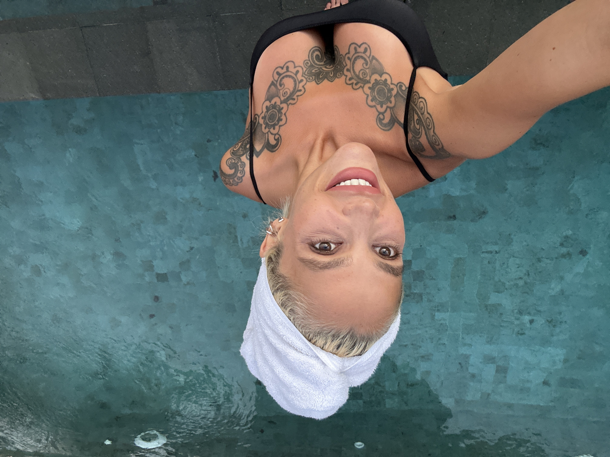 In Kuta Selatan, Indonesia, a woman with blonde hair wrapped in a white towel poses for a selfie by a pool. She is wearing a black swimsuit with thin straps, revealing tattoos on her chest and shoulders. The pool water is a vibrant turquoise color, and the background shows the tiled bottom of the pool. The woman is smiling, and her makeup is visible.