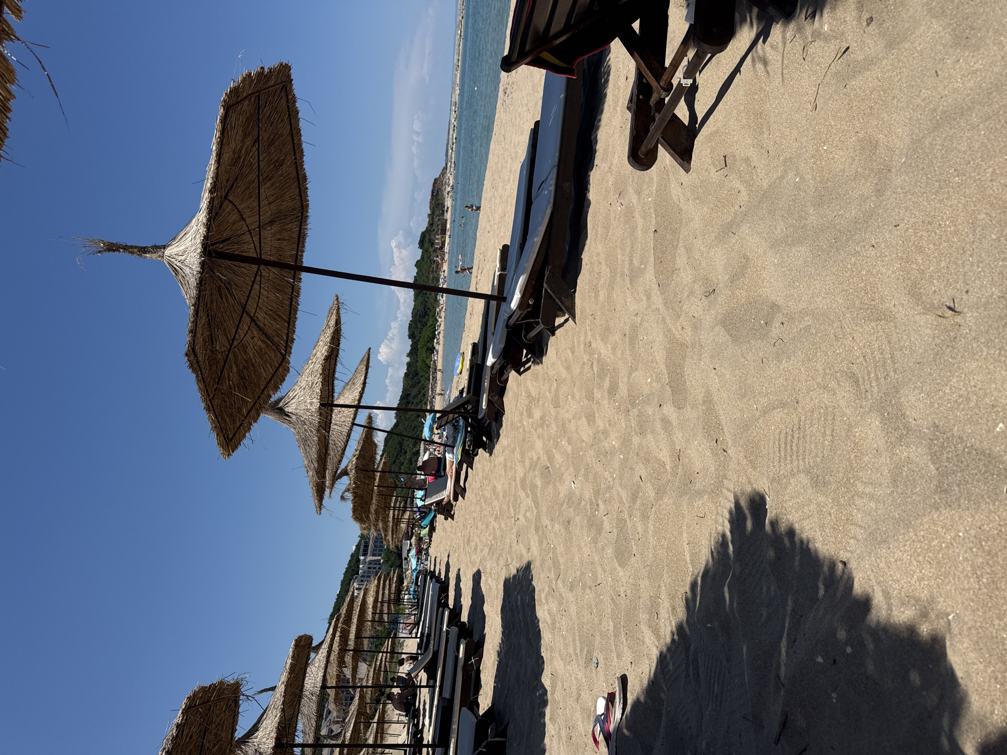 The image shows a sun-drenched beach in Dyuni, Bulgaria, with rows of unoccupied sun loungers and straw umbrellas set up on the sand. The calm sea is visible in the background, with a few people swimming and a distant coastline. The sand is light brown and appears well-maintained. The lighting suggests it is daytime, with clear blue skies and some scattered clouds. The foreground shows a pair of flip-flops and a shadow cast by one of the umbrellas, indicating a bright sunny day.