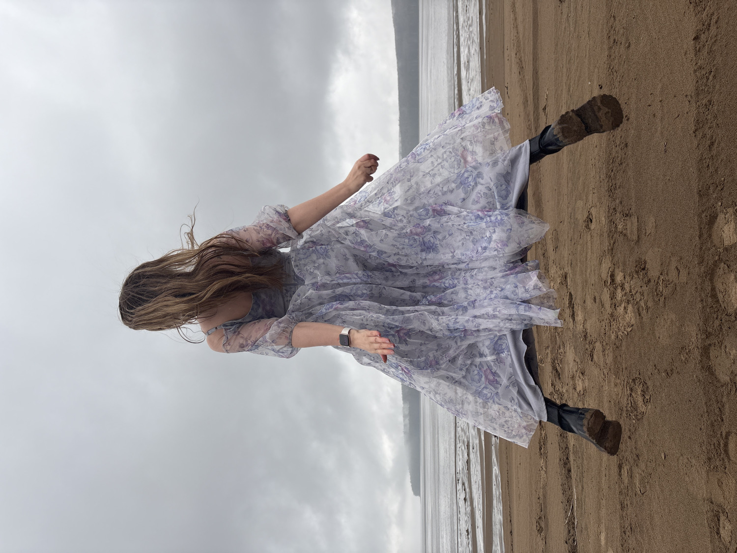 A woman is depicted from a slightly elevated perspective on a sandy beach in Chaikovsky, Russia, under an overcast sky. Her long, light brown hair flows in motion, partially obscuring her face as she appears to be turning or walking. She is wearing a light, sheer maxi dress with a delicate floral pattern in shades of purple and possibly green, layered over a solid grey slip. Dark, sturdy boots cover her feet, and a dark smartwatch is visible on her left wrist. Her arms are slightly outstretched, contributing to the sense of movement and the flowing appearance of her dress. The sandy beach extends towards calm water on the left side of the frame, with a grey, cloudy sky dominating the background. The scene suggests a cool, overcast daytime. There is no visible text in the image.
