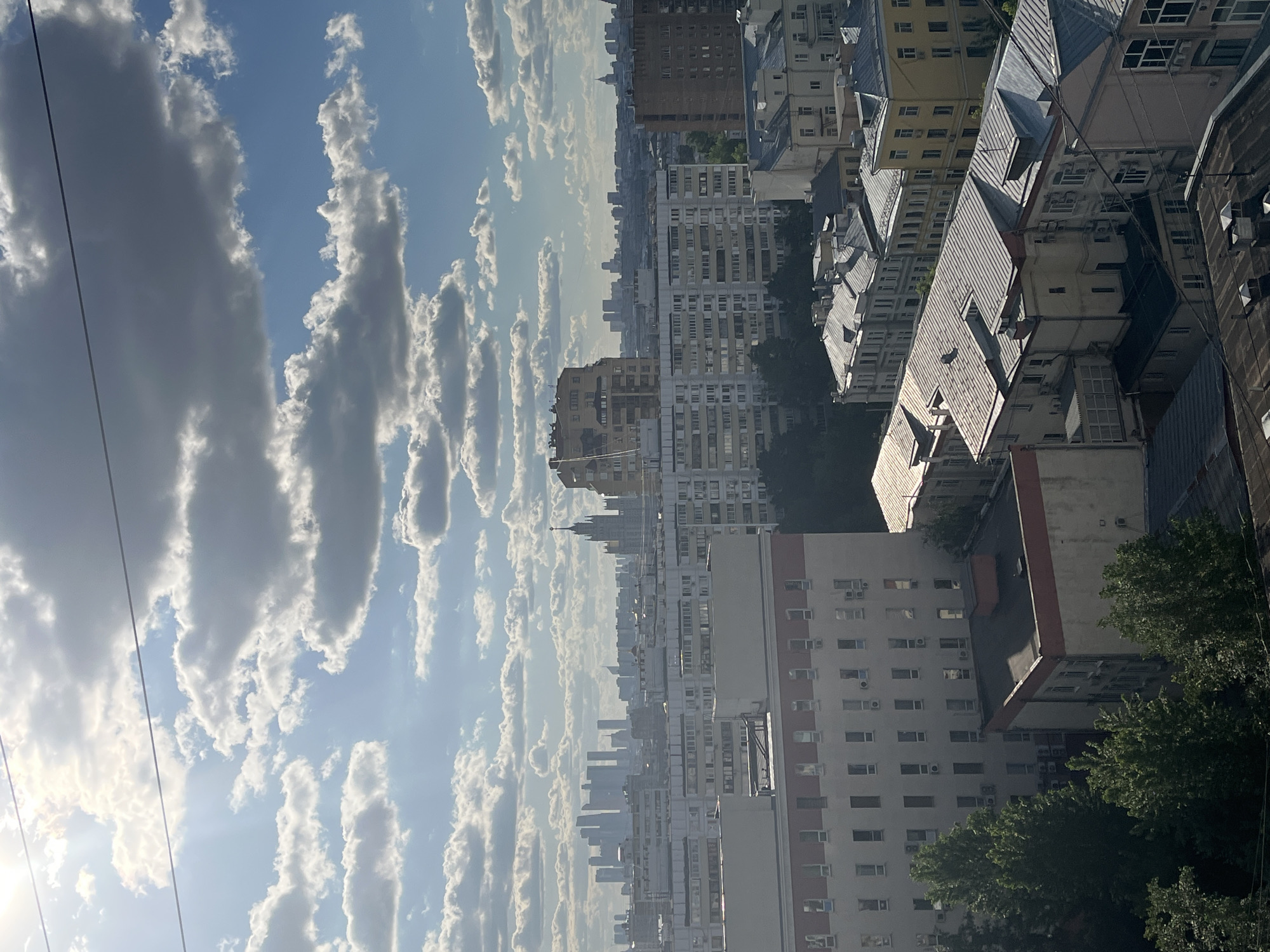 The image depicts an urban cityscape under a bright, partially cloudy sky. The perspective is from a high vantage point, likely a tall building or elevated area, looking towards a distant skyline. In the foreground and midground, there are numerous buildings of varying heights and architectural styles, with a mix of residential and commercial structures. The buildings closest to the viewer have visible details, such as windows, rooftops, and some greenery around their bases. In the distance, the city skyline is more obscured by atmospheric haze, but it still shows a dense arrangement of taller buildings. The sky is filled with white, fluffy clouds, contrasting with the blue sky in between. There are also some visible utility wires cutting across the sky in the foreground.