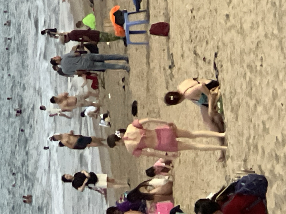 The image presents a crowded beach scene in Nha Trang, Vietnam. The sandy beach occupies the foreground and midground, transitioning to the ocean in the background. The water shows moderate waves with visible whitecaps, populated by approximately 20-30 individuals swimming, wading, and standing.
In the immediate foreground, a person wearing a red top and a blue backpack is partially visible on the far left. Centrally, a female stands with her back to the viewer, wearing a pink off-shoulder two-piece swimsuit. To her right, another female is seated on the sand, wearing a dark blue and light blue bikini top with dark bottoms.
In the midground, a female in a black top and white skirt stands facing the water. Behind her, a group includes a woman in a purple top and a child in pink crawling on the sand near a pink bag. Further right, two men are standing and conversing; one wears a light blue shirt and jeans, the other a dark red shirt, camouflage shorts, and a cap. Nearby, an orange fabric item and a dark red bag are on the sand, beside a blue plastic stool. Several shirtless men are visible closer to the water's edge, wearing shorts and engaged in wading or entering/exiting the water. A person in a bright yellow-green shirt is seated further back in the midground right. Scattered footwear and unidentifiable small objects are present on the sand throughout the midground.