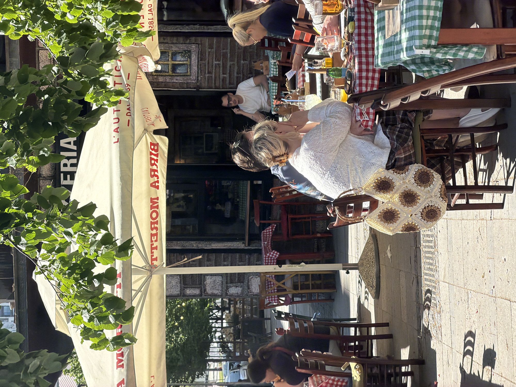 The image depicts an outdoor scene in Belgrade, Serbia. Several people are seated at tables outside a restaurant, enjoying food and drinks. The restaurant has a large awning with the word "BIRRA" prominently displayed. The tables are covered with red and white checkered tablecloths and green and white checkered tablecloths. Some patrons are engaged in conversation, while others are looking at menus or interacting with food. The setting appears to be a sunny day. The building behind the restaurant has a brick facade. There are trees and plants visible in the foreground, adding to the outdoor ambiance. The seating area extends onto a stone-paved sidewalk.