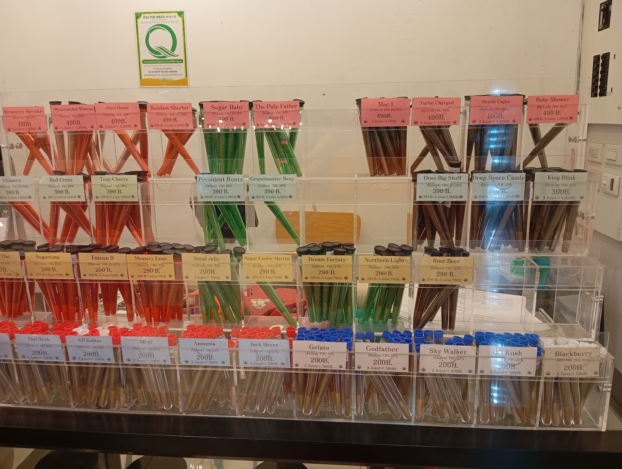 The image depicts an interior view of a cannabis shop in Karon, Thailand. The focus is on a multi-tiered display case filled with pre-rolled cannabis joints packaged in clear plastic tubes. Each tube is labeled with the strain name, THC percentage, price, and a quantity deal. The display is organized into rows and columns, showcasing a variety of strains with different colors of packaging and joint contents. The background reveals the shop's branding, including a sign with "THE WEED SHOP 2" and a circular green logo. The lighting is bright, illuminating the products and the surrounding environment, which appears to be a retail space. No people are visible in the image.