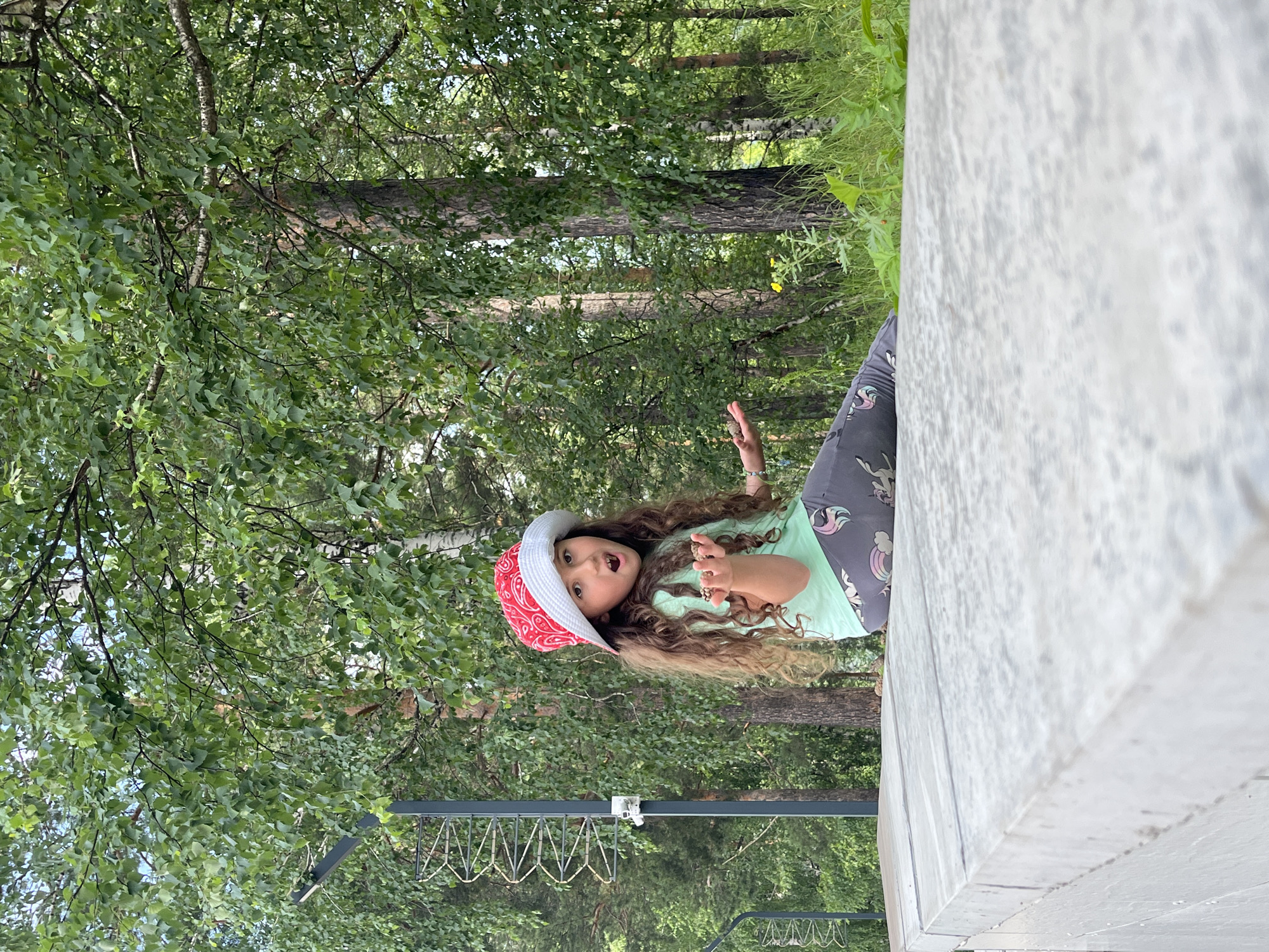 A young girl with long, curly brown hair is sitting on a light-colored stone ledge in front of a forest. She is wearing a red and white patterned hat, a light green t-shirt, and grey leggings with a unicorn print. Her mouth is open, and she is holding a small handful of something in her right hand, which is extended. Her left hand is resting on her leg. Behind her, there are tall trees with green leaves, and the ground is covered with grass. A dark pole with a coiled wire structure is visible to the left of the girl. The scene appears to be outdoors in a natural setting, likely a park or wooded area. The location is the city of Divnogorsk, Russia.