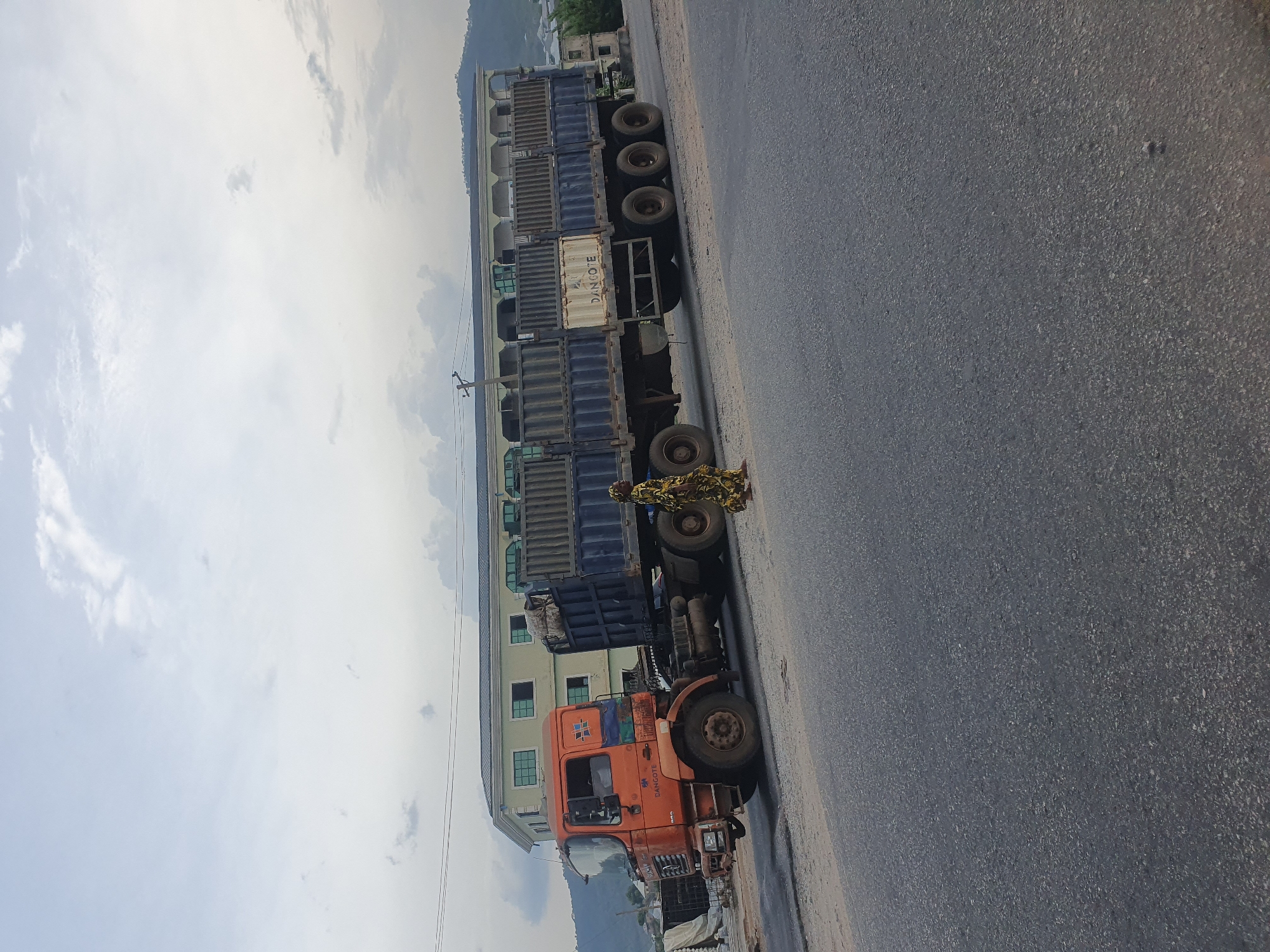 The image displays a roadside scene in Lokoja, Nigeria, under a bright, partly cloudy daytime sky. An orange semi-truck is prominently featured on the right side of an asphalt road, appearing to be parked or stopped. The truck's cab is orange, showing signs of wear, with blue and white accents. The word "DANGOTE" is clearly visible in white lettering on the side of the truck's cab.
Attached to the truck is a long trailer loaded with multiple cargo containers. Most containers are dark blue, but one white container, also bearing the "DANGOTE" logo, is visible towards the front of the trailer.
A person dressed in distinctive patterned clothing, primarily yellow and black, stands next to the truck's trailer, near the rear wheels of the cab.
In the background, a long, multi-story building with a light-colored facade and numerous windows stretches along the road. Beyond the building, distant hills or mountains can be seen under the expansive sky. A utility pole with wires is also present on the left side of the road, close to the foreground. The road itself is made of dark asphalt.
The presence of the large commercial truck and containers, coupled with the "DANGOTE" branding, suggests a setting related to freight transport and industry.
