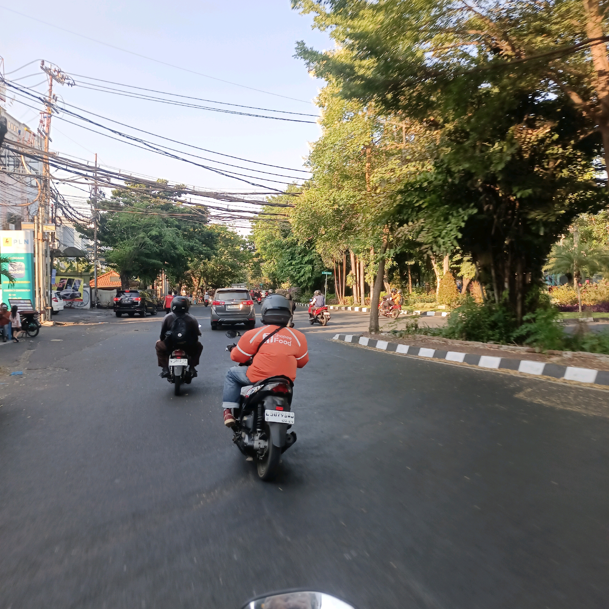 This is a street-level, first-person perspective photograph taken on a paved road in Surabaya, Indonesia, possibly during late afternoon given the lighting. The road is wide and appears to be a multi-lane street. On the right side of the road, there is a park-like area with a variety of trees, including large, mature trees with dense foliage and some palm trees, bordered by a sidewalk with a distinctive black and white striped curb. On the left side of the road, there are buildings with a significant amount of overhead electrical and telecommunication wires, suggesting an urban environment.
Several motorcycles are present. In the foreground, a person is riding a motorcycle with an orange jacket that has "Shopee Food" written on the back. This individual is wearing a helmet and blue jeans. Another motorcyclist in front is wearing a black jacket and a helmet. Further down the road, other motorcycles and a few cars are visible. One car, a black SUV, is in the middle lane. The sky is clear and blue, indicating good weather. The overall impression is of a busy urban street with heavy vehicular traffic and a mix of commercial and green spaces.