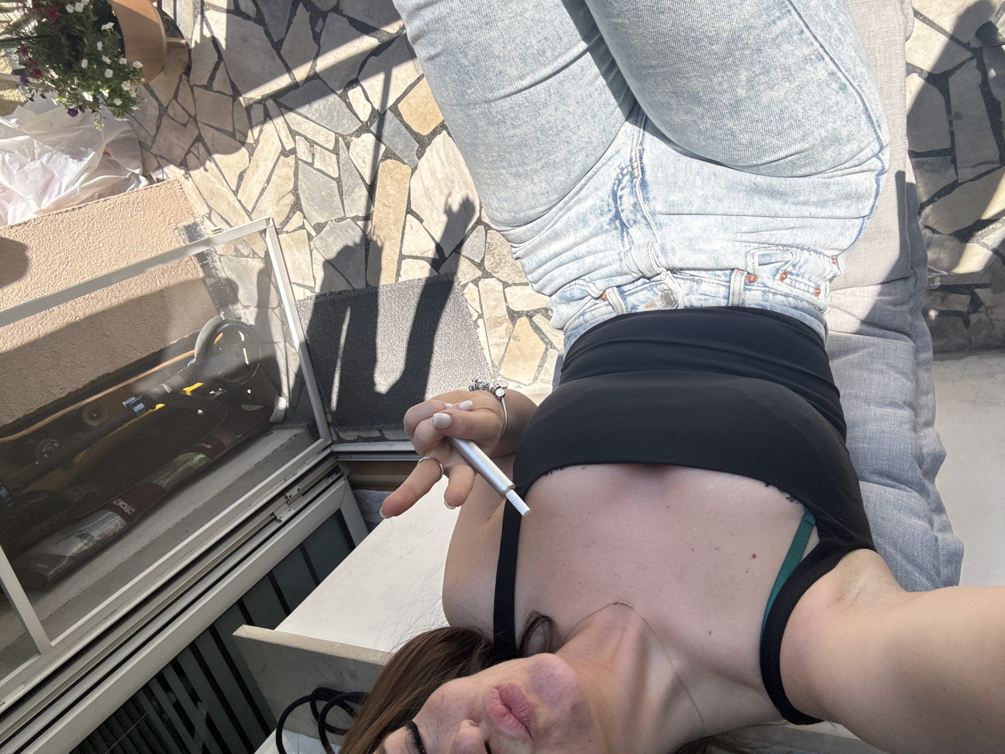 A woman lies on a gray, cushioned outdoor sofa on a stone tile patio. She wears light blue jeans and a black top, and holds a silver vaping device. A window reflects skateboards. The background shows flowers in a pot and glimpses of Plovdiv.