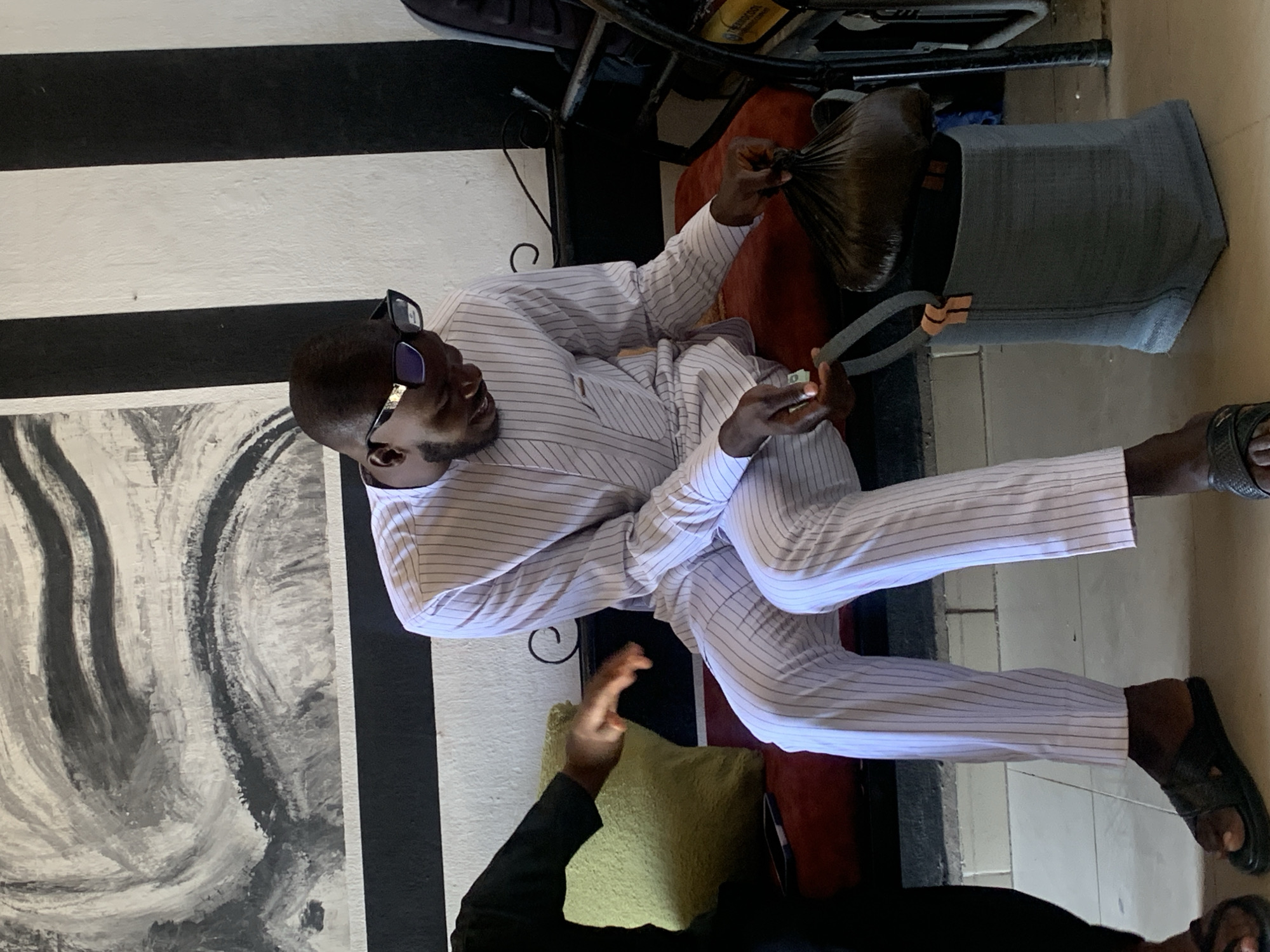 A man wearing a light-colored striped suit and sunglasses is seated indoors. He holds a dark plastic bag in his left hand and what appears to be a folded bill in his right. He also has a pair of sandals on his feet. Next to him, there is a gray bag and a green pillow. Another person is partially visible at the bottom of the image. A black and white striped wall is in the background, adjacent to an abstract painting or drawing. The location is likely in Jos, Nigeria, given the prompt's instruction.