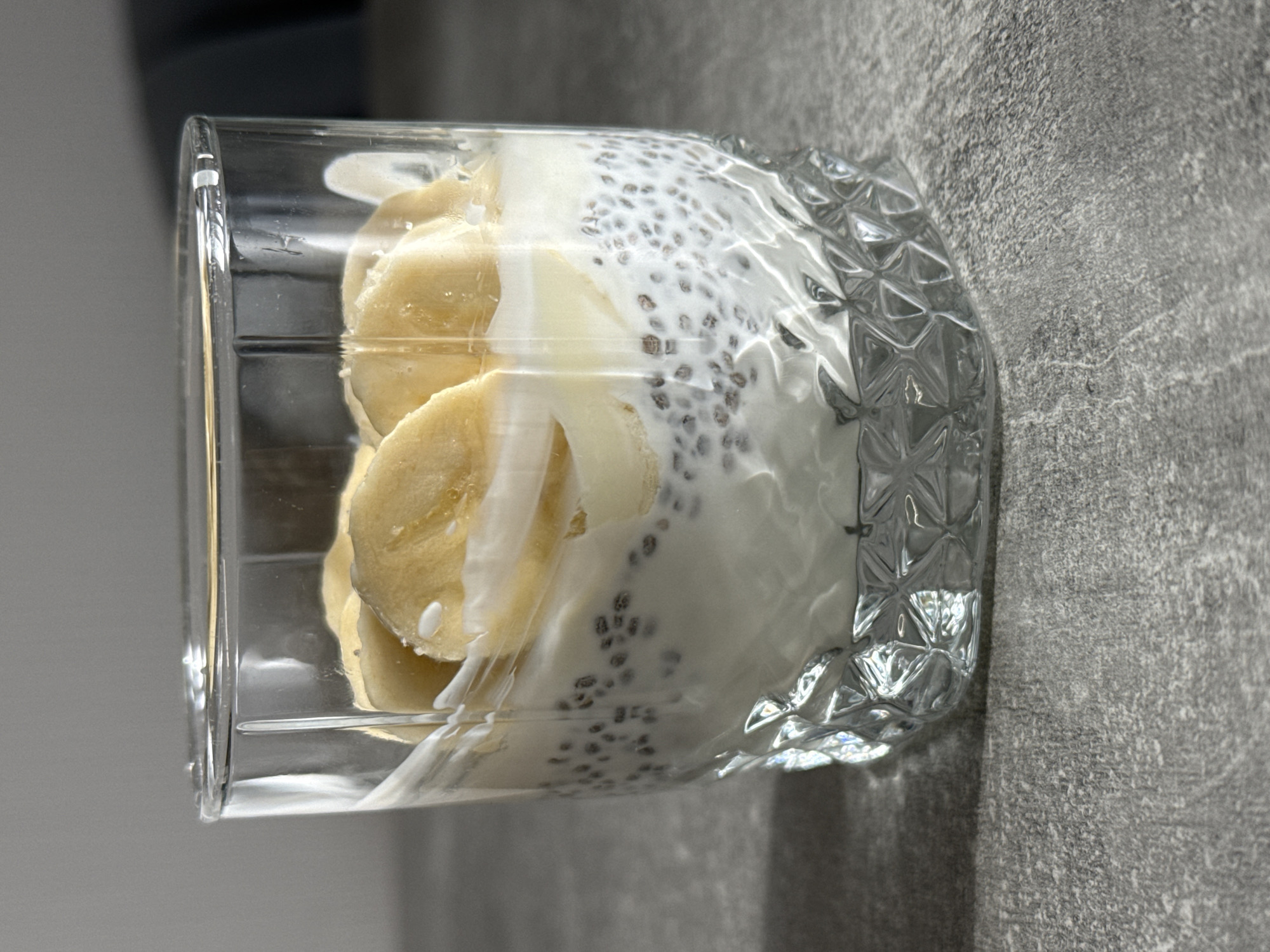A clear glass tumbler, oriented horizontally as if lying on its side, is filled with a layered food item against a textured grey background. The contents of the glass include several slices of light yellow banana and a creamy white substance, likely yogurt or milk, mixed with numerous small, dark chia seeds. The chia seeds are visibly dispersed throughout the white base. The glass itself is transparent with a smooth rim, and its base features a decorative, faceted or patterned texture that reflects light. The background is a solid, medium grey surface with a rough, mottled texture, possibly a wall or a countertop. There are no people or discernible activities taking place. No text is visible. The lighting is soft and even, highlighting the details of the food and the glass.
