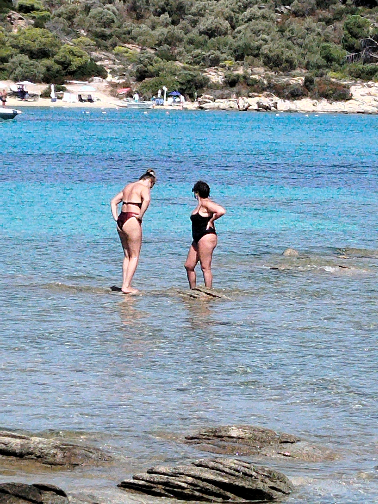 Two women stand in shallow, clear turquoise water near a rocky shoreline. The woman on the left wears a maroon bikini and faces away from the viewer, looking down. Her hair is pulled up in a bun. The woman on the right wears a black one-piece swimsuit and faces left, with her right hand on her hip. Her dark hair is short. In the background, a sandy beach is visible with trees and vegetation on a hillside above. Several beach umbrellas and loungers are present on the beach, along with a small boat in the far left of the frame. The water is calm with ripples and the seabed is visible in places.