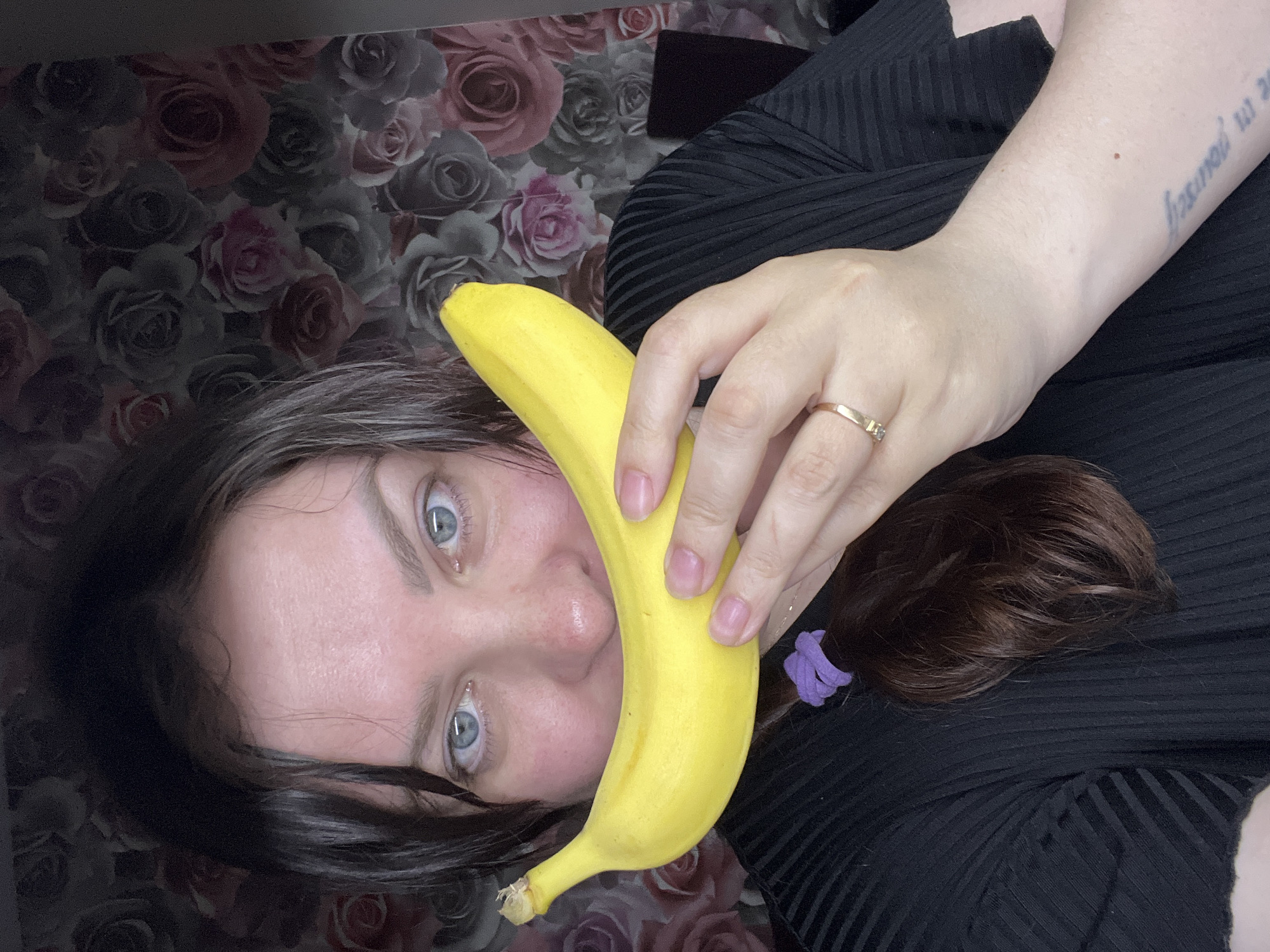 A woman with blue eyes and brown hair holds a yellow banana in front of her face. She is wearing a black top and a ring on her finger. The background is a floral pattern. The location is not discernible.