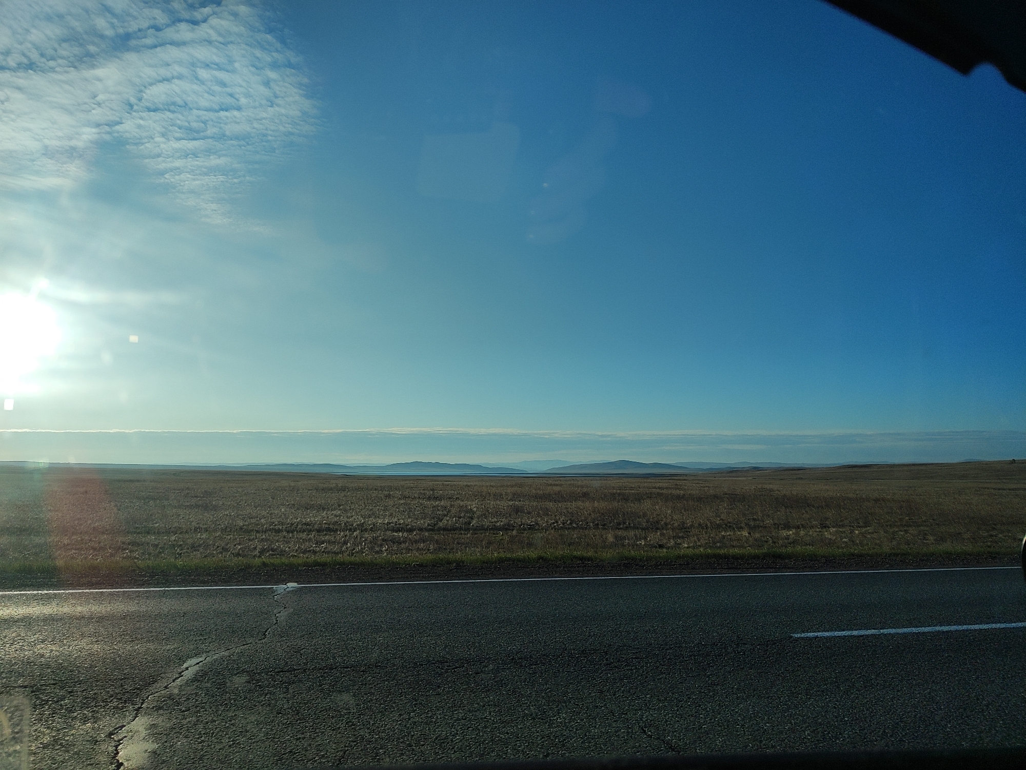 The image depicts a landscape viewed from inside a vehicle, likely a car. The foreground consists of an asphalt road with white lane markings and cracks. Beyond the road, a field of dry, brown vegetation stretches to the horizon. In the distance, a faint line of hills or mountains can be seen under a clear, blue sky with some wispy clouds. The sun is visible on the left side of the frame, creating a bright glare. The location appears to be a rural or sparsely populated area. The specific city is unknown.