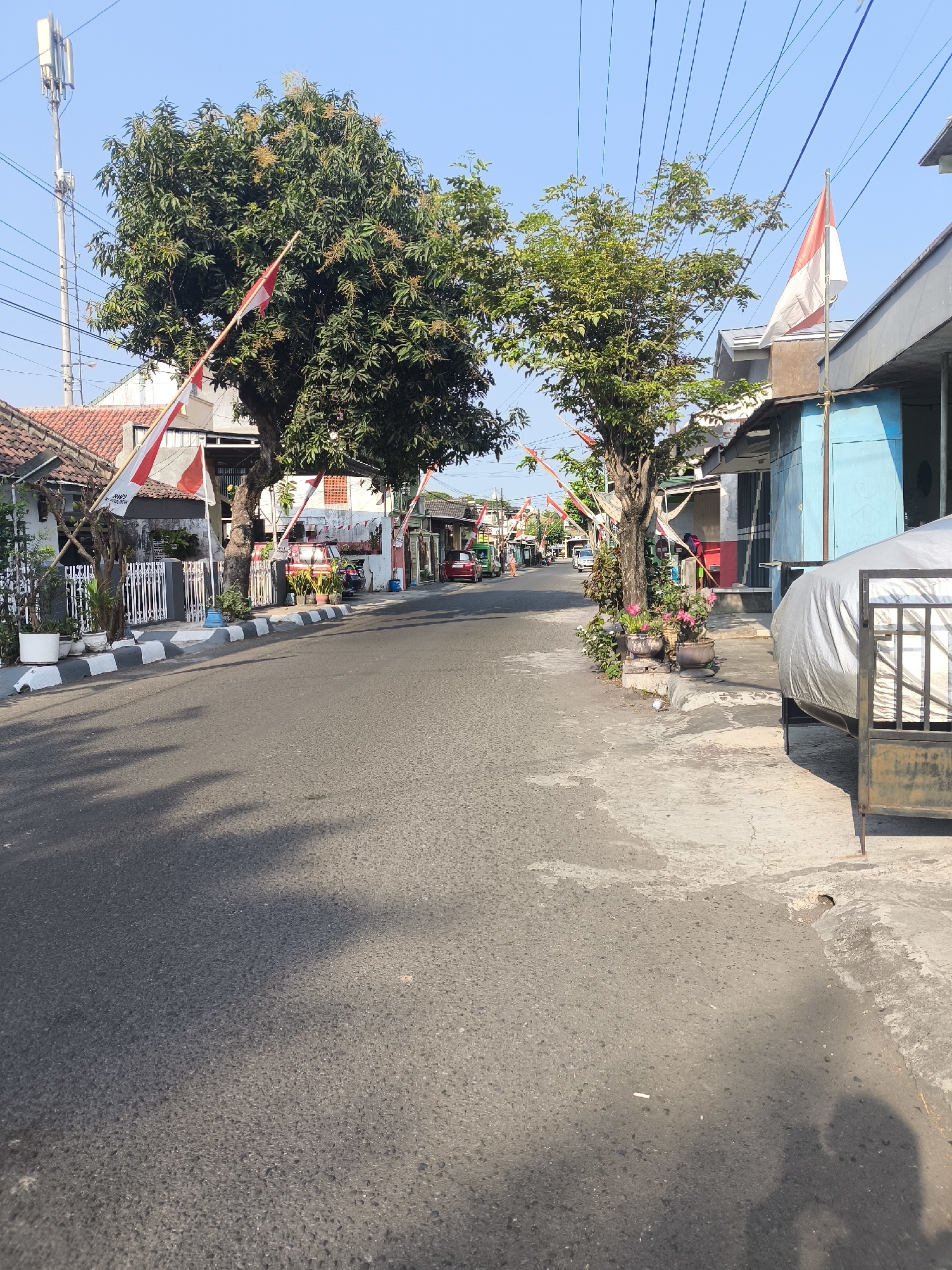 The image depicts a street scene in the city of Citrodiwangsan, Indonesia, likely on a bright, sunny day. The road surface is asphalt, with a distinct black and white striped curb along the left side. Houses with red-tiled roofs and white fences line the street, decorated with Indonesian flags and pennants, suggesting a national celebration or commemoration. Trees with green foliage are visible, some bearing what appear to be developing fruits. Power lines crisscross overhead against a clear blue sky. Several parked vehicles, including a red car and a green van, are present. On the right side of the road, a blue-fronted building stands next to a structure covered with a silver tarp and enclosed by a metal gate. The overall impression is of a residential street adorned for a special occasion.