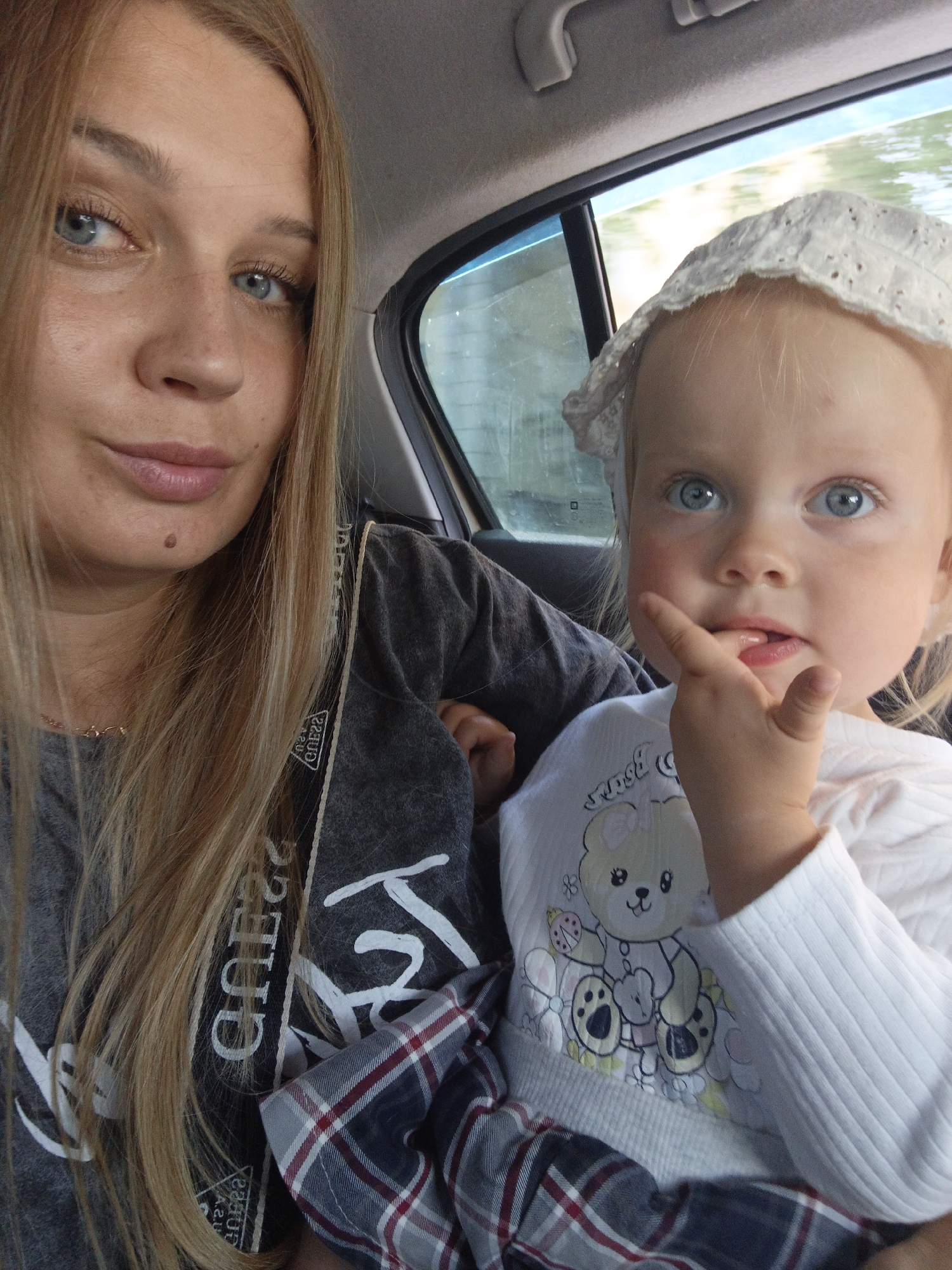 A woman with blonde hair and blue eyes is in the passenger seat of a car, taking a selfie with a toddler in her arms. The toddler, also with blue eyes, is wearing a white bonnet and a white shirt with a teddy bear print. The woman is wearing a grey t-shirt with a "Guess" logo visible on it. The interior of the car is visible, with the window showing a blurred background of trees and sky, suggesting the car is in motion. The scene is lit by natural daylight.
